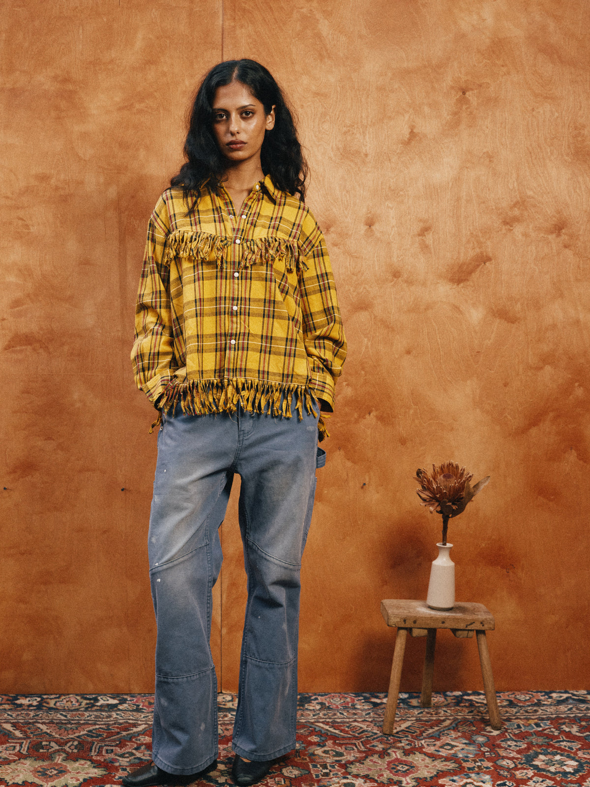 A woman leans against a wooden wall in the High Plains Fringe Shirt by FOUND and loose jeans. Nearby, a stool with a vase and dried flower rests on a patterned rug.