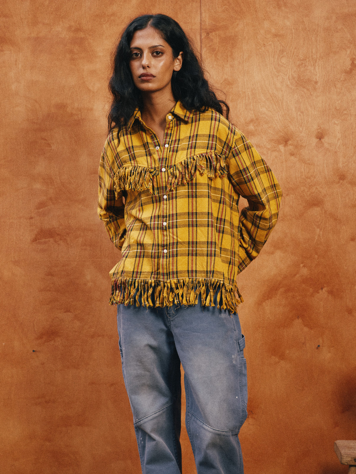 A person with long dark hair wears the FOUND High Plains Fringe Shirt in yellow plaid and blue jeans, standing against a brown textured backdrop.