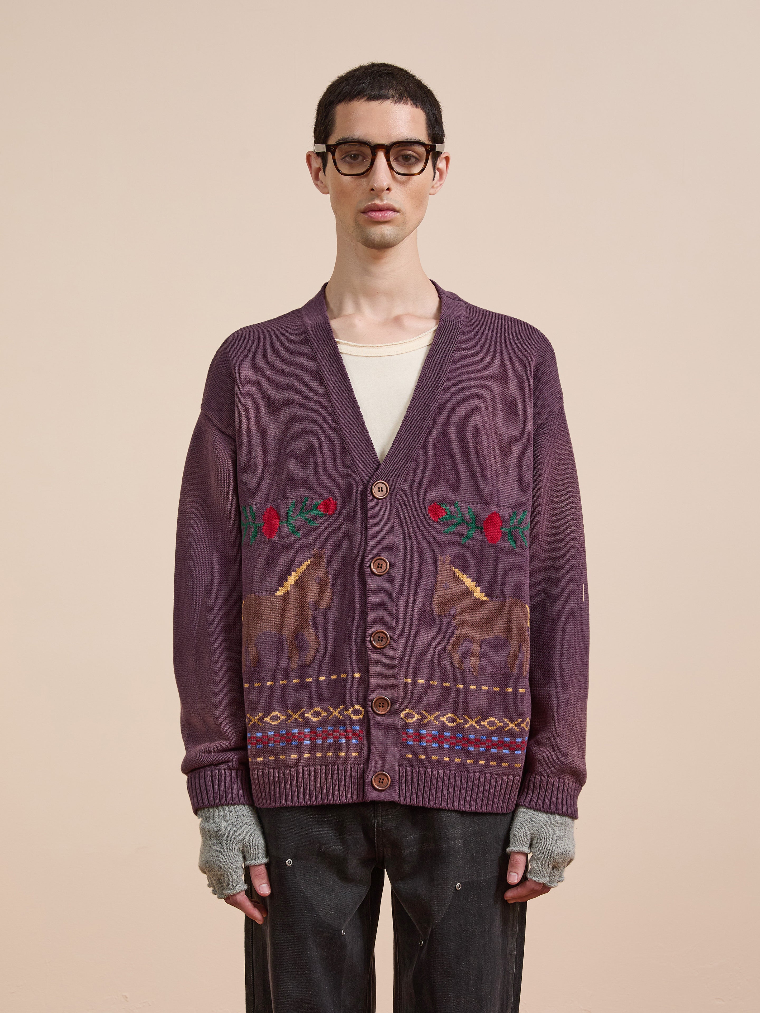 A person in glasses, a white shirt, dark pants, and fingerless gloves wears the Found Horse Jacquard Cardigan with floral and horse motifs, standing against a plain beige background.