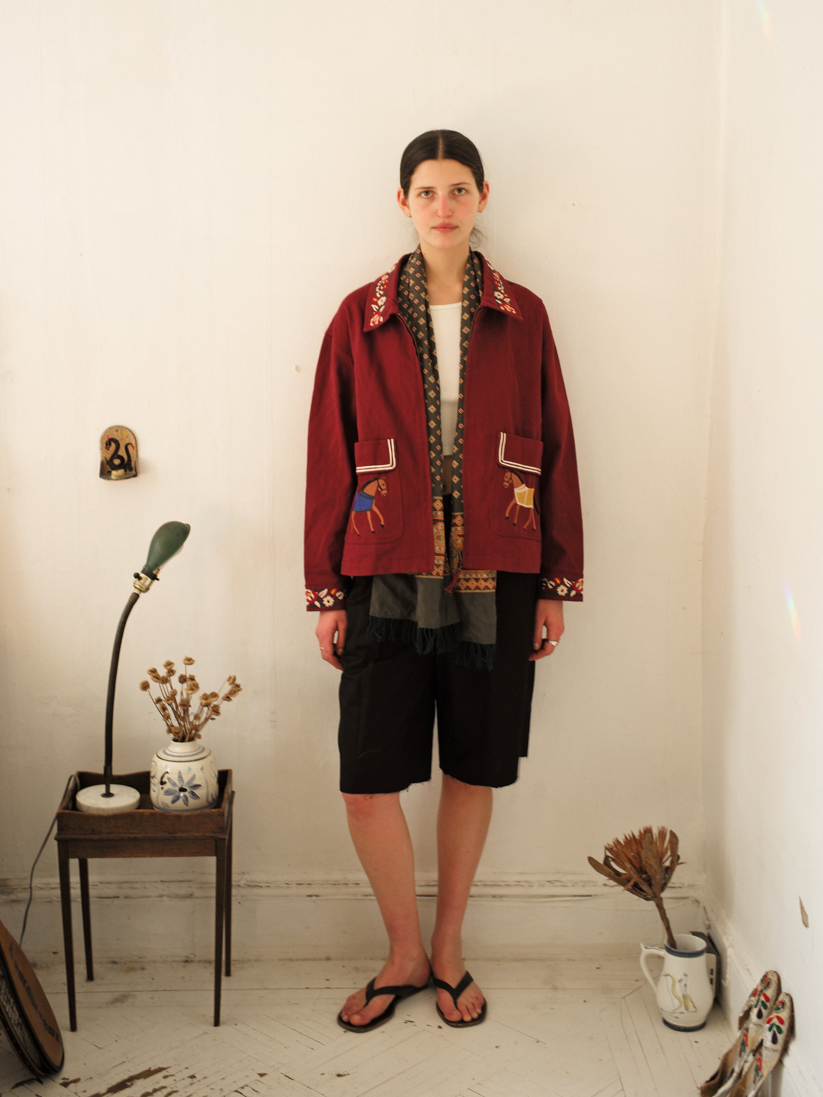 A person wearing the Oxblood Horse Equine Jacket by FOUND over a white shirt and black shorts stands in a room with white walls, next to a small table adorned with a lamp and dried flowers.