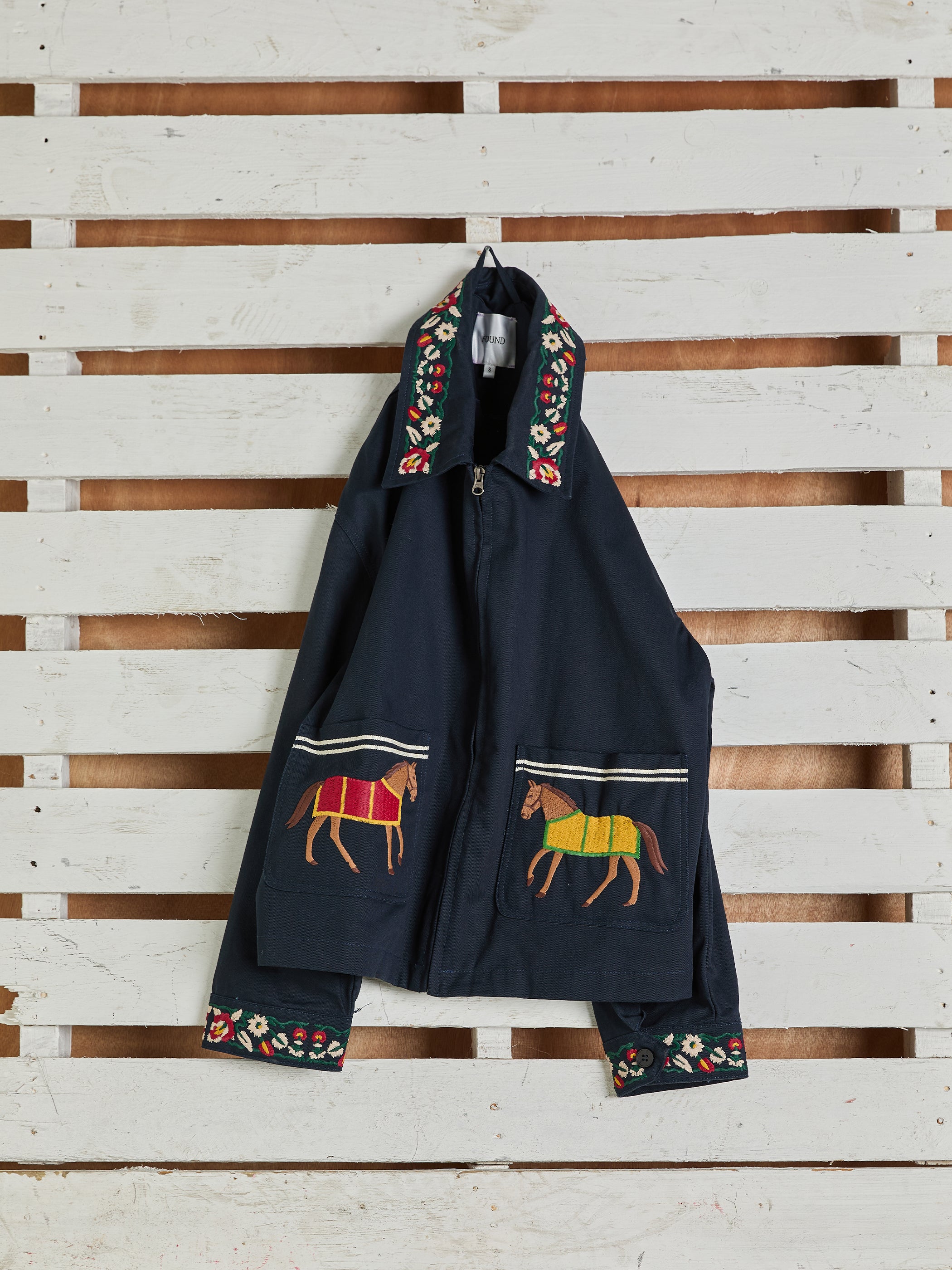 The Horse Equine Work Jacket by Found, inspired by 70's jockey uniforms, showcases exquisite horse embroidery on the front and vibrant floral patterns on the collar and cuffs. It's crafted from durable cotton twill fabric and elegantly displayed against a white wooden backdrop.