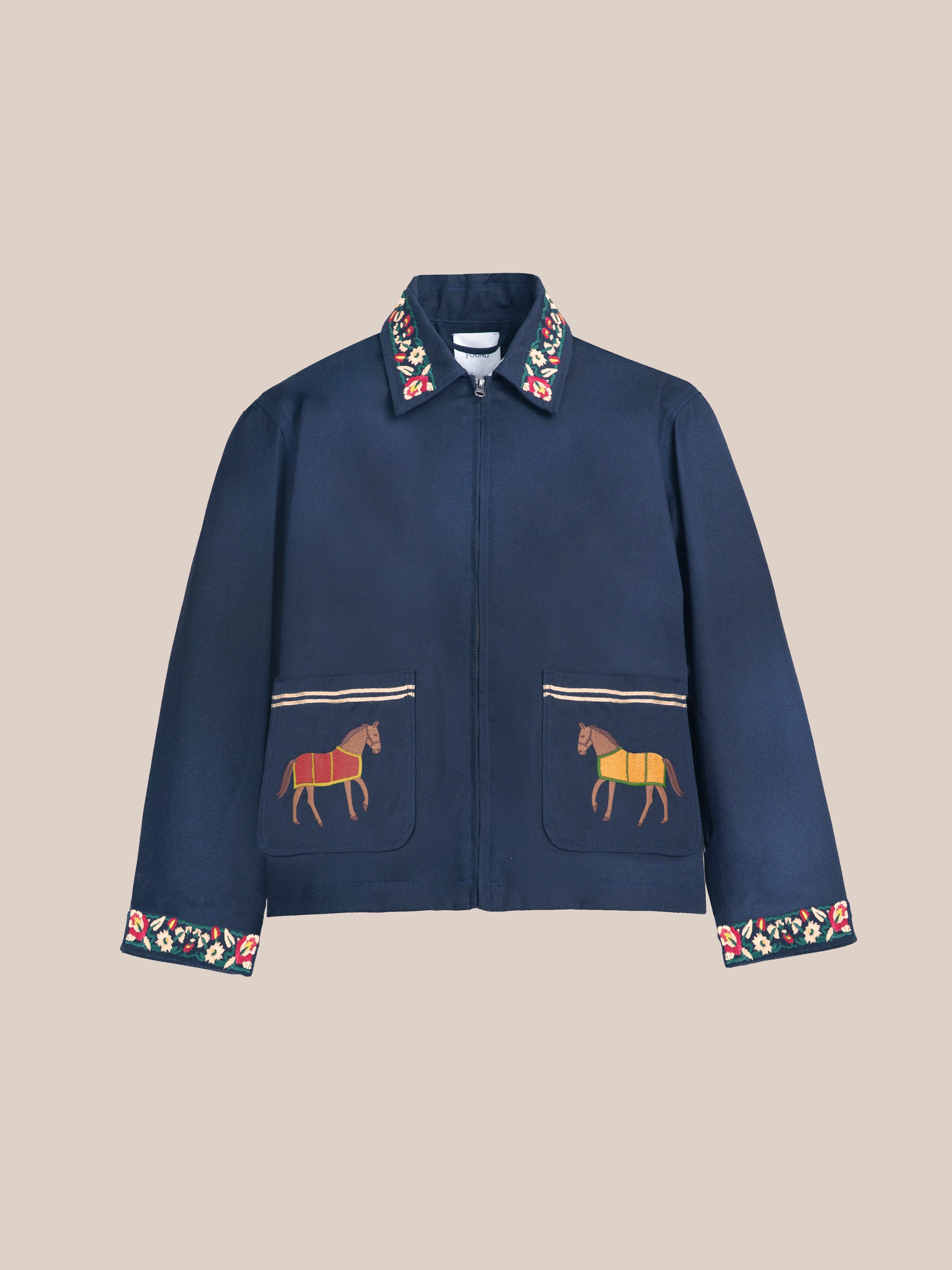 The Found Horse Equine Work Jacket boasts floral embroidery on the collar and cuffs, features horse designs on the front pockets, and showcases subtle color blocking against a beige background in navy blue.