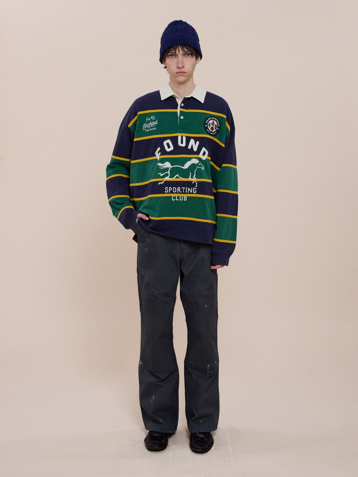 Person wearing a blue knit beanie, FOUND Horse Logo Rugby, loose dark pants, and black shoes stands against a plain beige background.