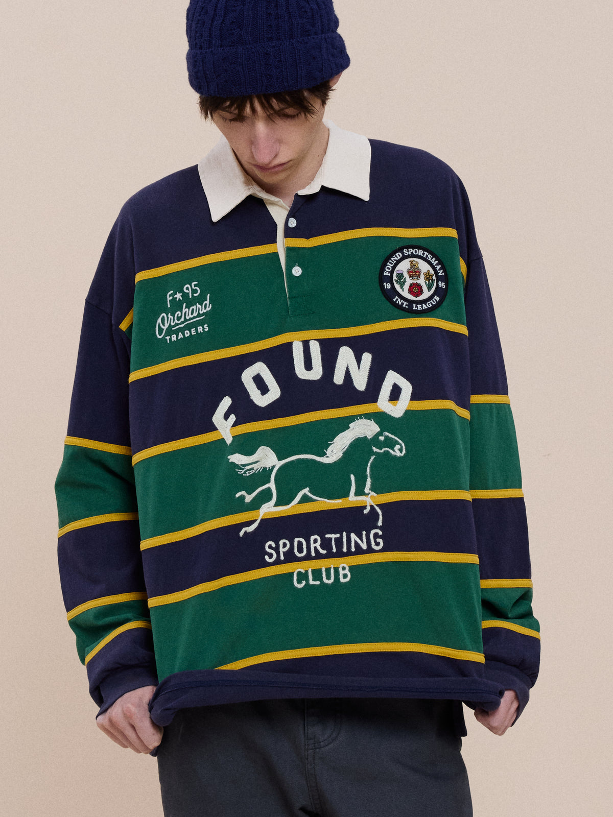 A person in a navy blue knit hat and the Horse Logo Rugby by FOUND with “FOUND SPORTING CLUB” on the front looks down.