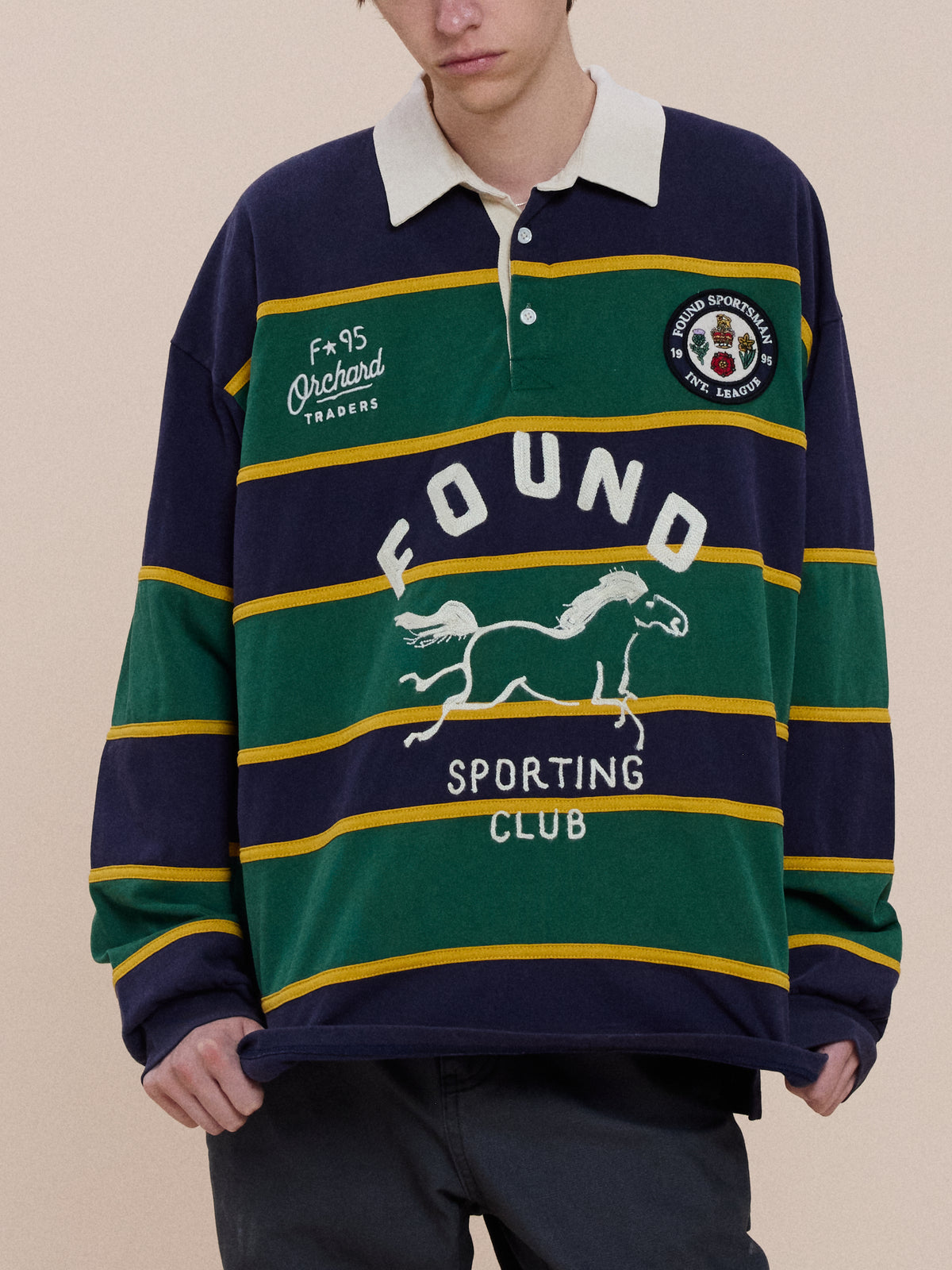 Person wearing the FOUND Horse Logo Rugby, a unisex shirt with navy, green, and yellow stripes, white collar, horse graphic, and “FOUND SPORTING CLUB” on front.