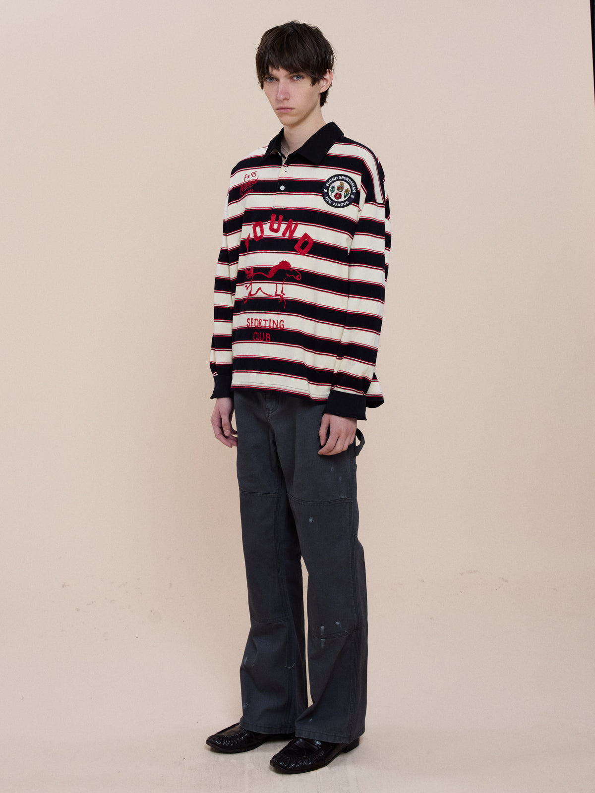 A person stands against a plain background, wearing the FOUND Horse Logo Rugby, dark trousers, and black shoes.