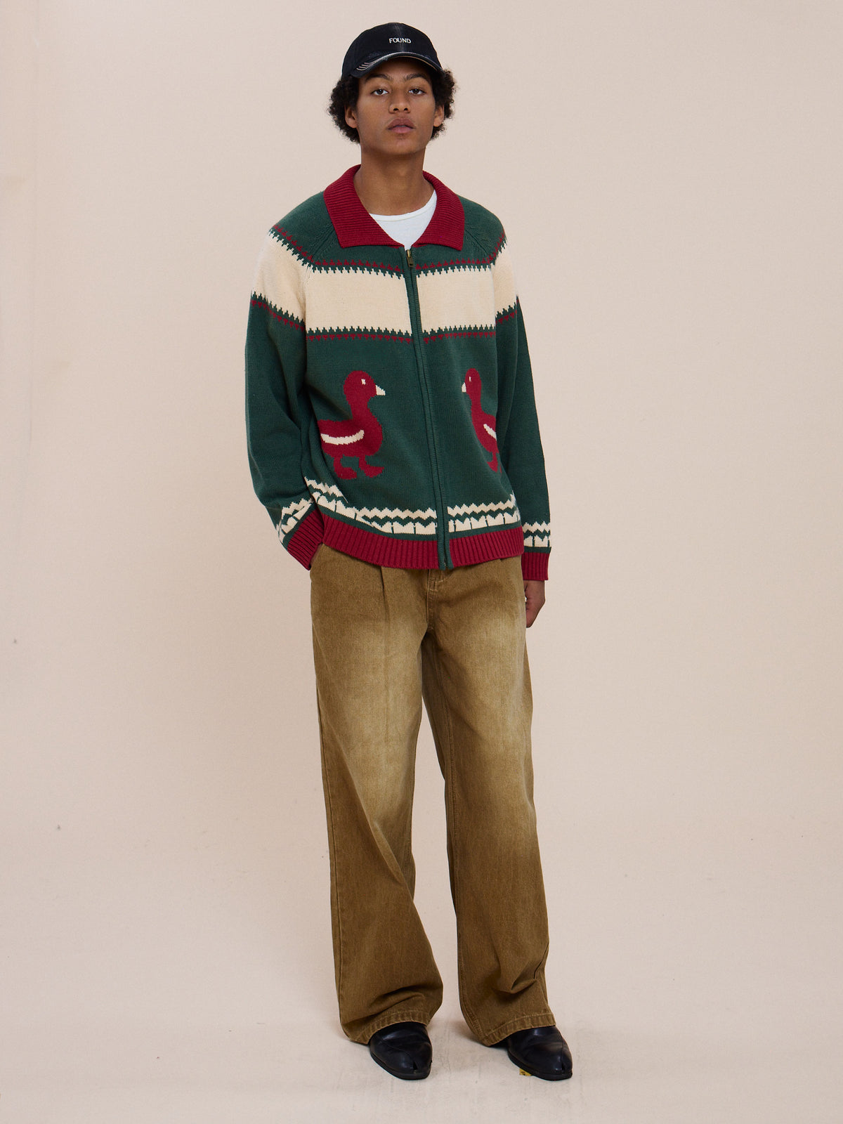 Person wears a black cap, Isles Sweater by FOUND, wide brown pants, and black shoes while standing against a plain beige background.