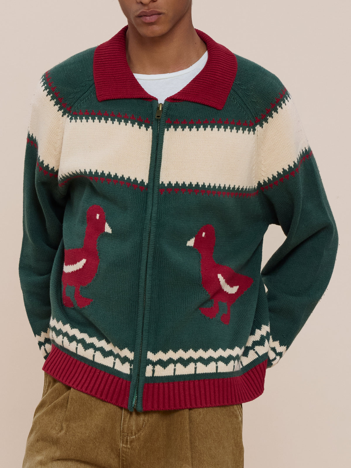 A person styles the FOUND Isles Sweater—green with red ducks, cream/red patterns, and a red collar—with tan corduroy pants for a vintage knitwear look.