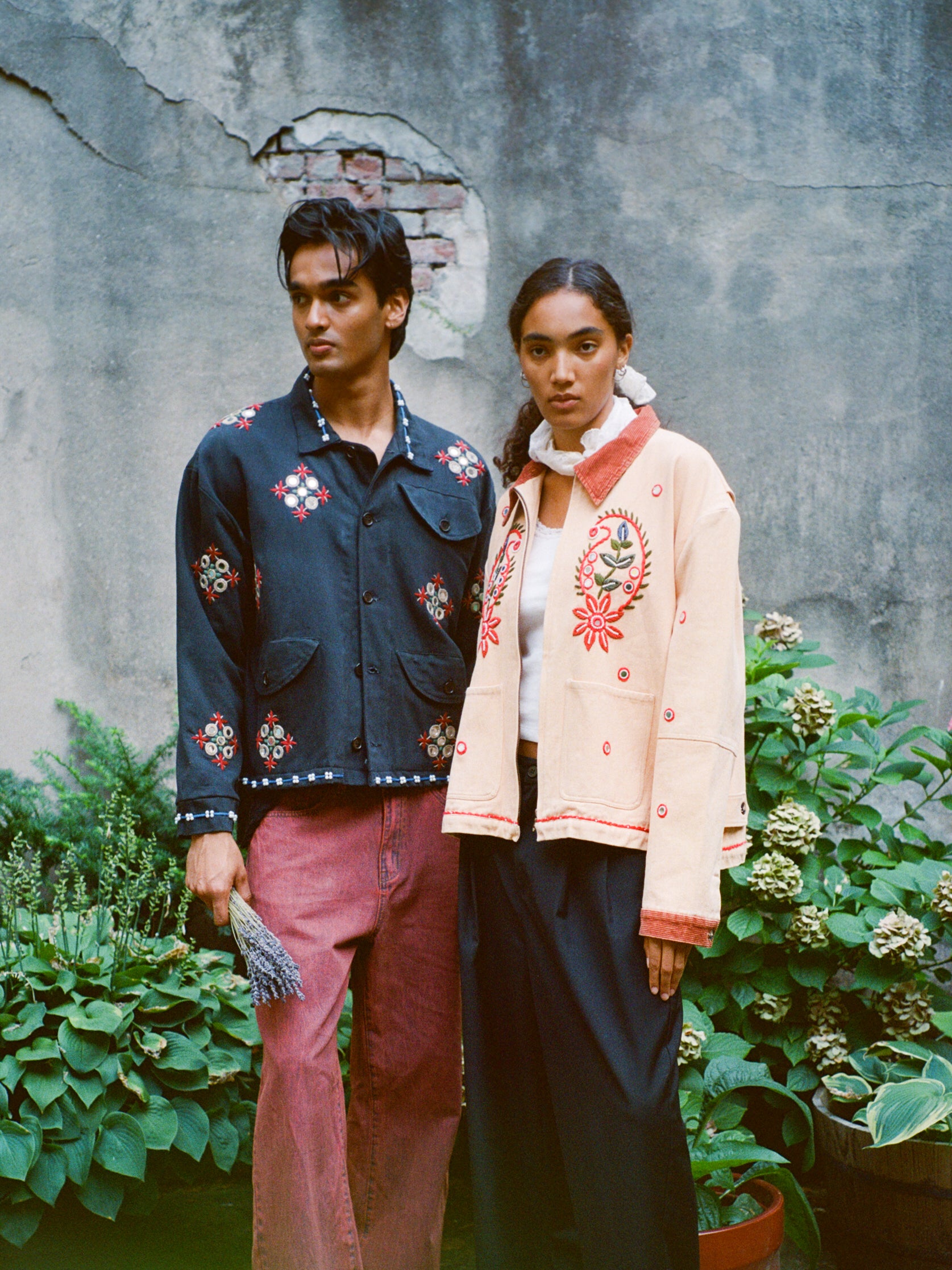 Two people stand before a weathered wall and plants, wearing Found's Mirror Ornament Hand Embroidered Work Jacket. The cotton twill jackets feature intricate Aari Sheeshakari embroidery. One holds dried flowers; both have neutral expressions.