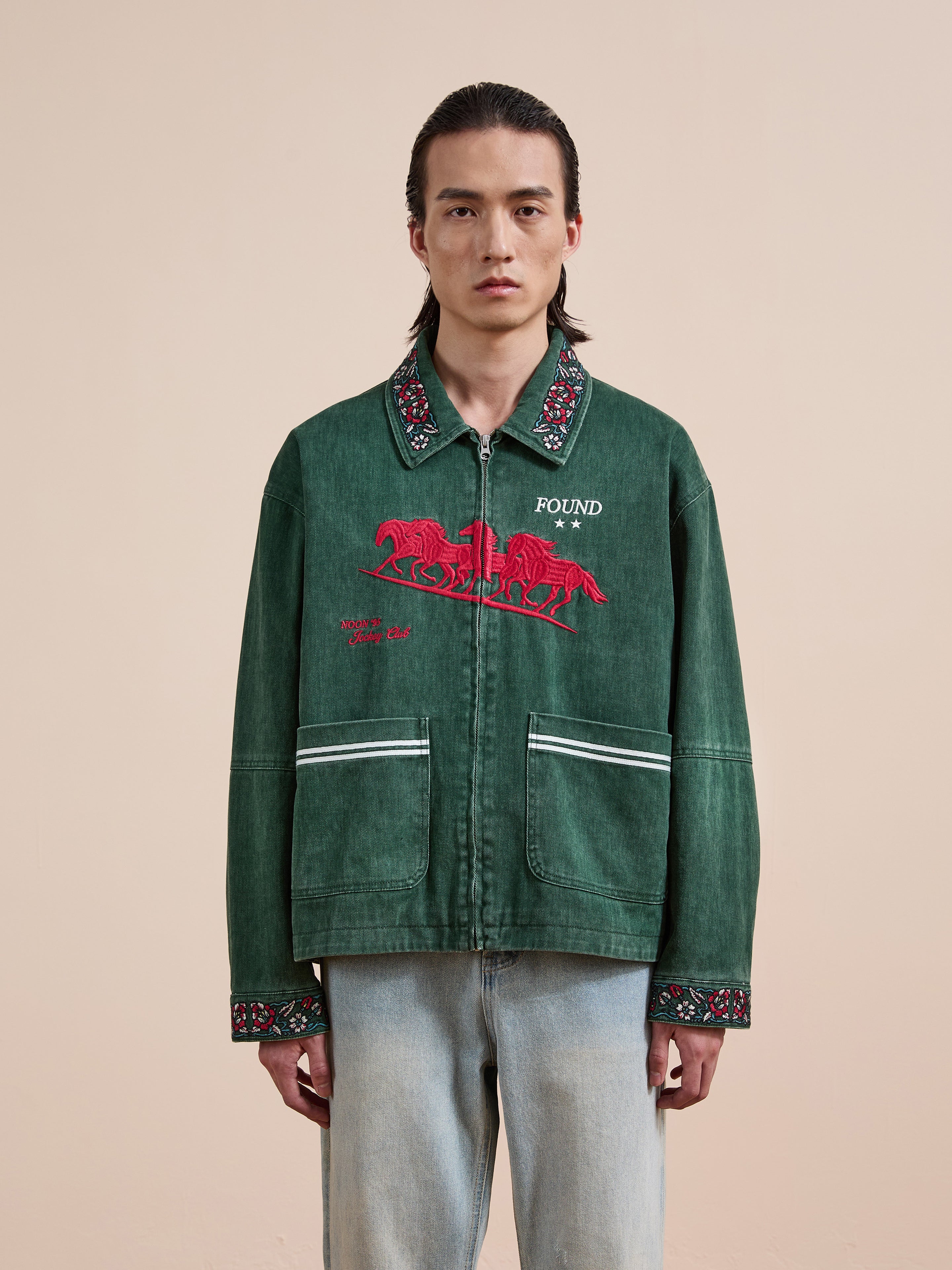 A person wears the Found Jockey Club Embroidered Jacket—green with racing horses, floral-trimmed collar and cuffs—and light blue jeans, standing against a beige background. This unisex relaxed fit look is from Found.