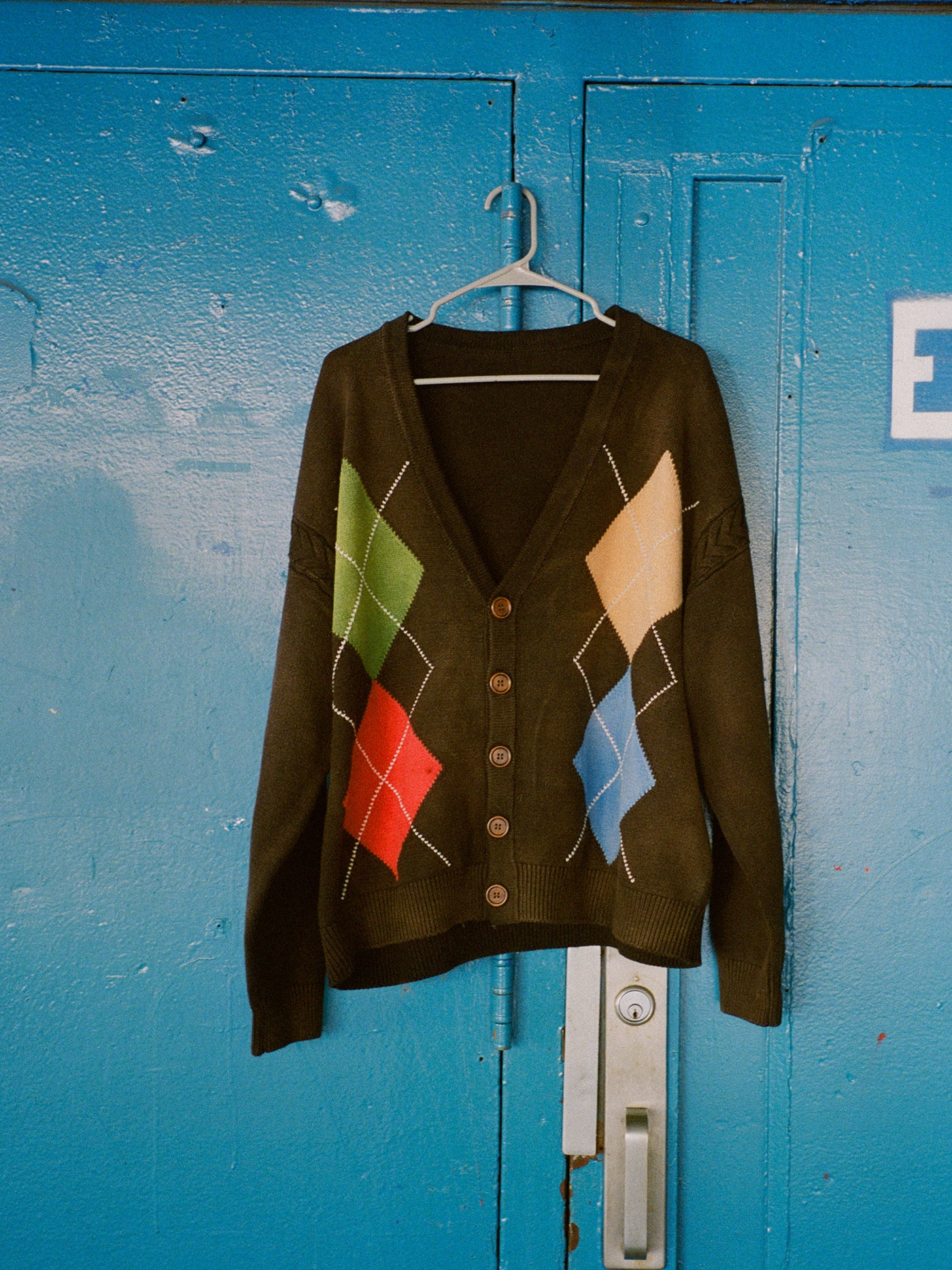 The FOUND Argyle Knit Cardigan, featuring black fabric with red, green, yellow, and blue diamond patterns, hangs on a plastic hanger against a blue metal door.