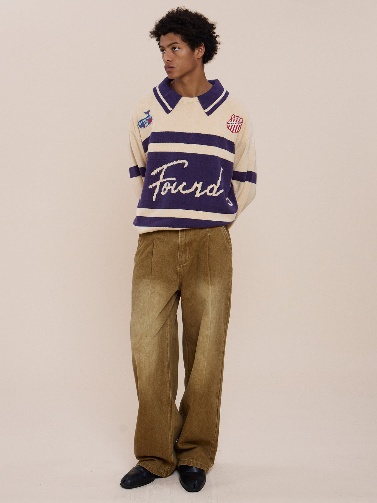 A person wears FOUND’s Knit Jersey in cream and purple, paired with loose tan pants and black shoes, standing against a plain beige background, gazing to the side.