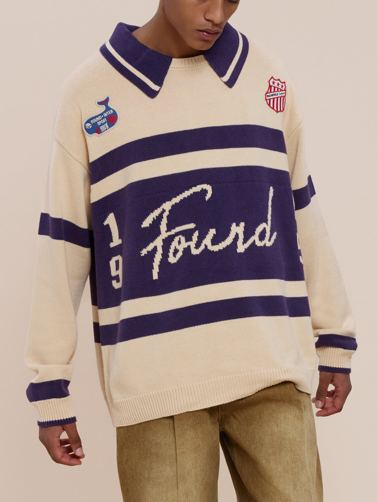 Person wearing a beige and purple FOUND Knit Jersey with “Found” on the front, “1949” on the sides, two chest patches, paired with tan pants.