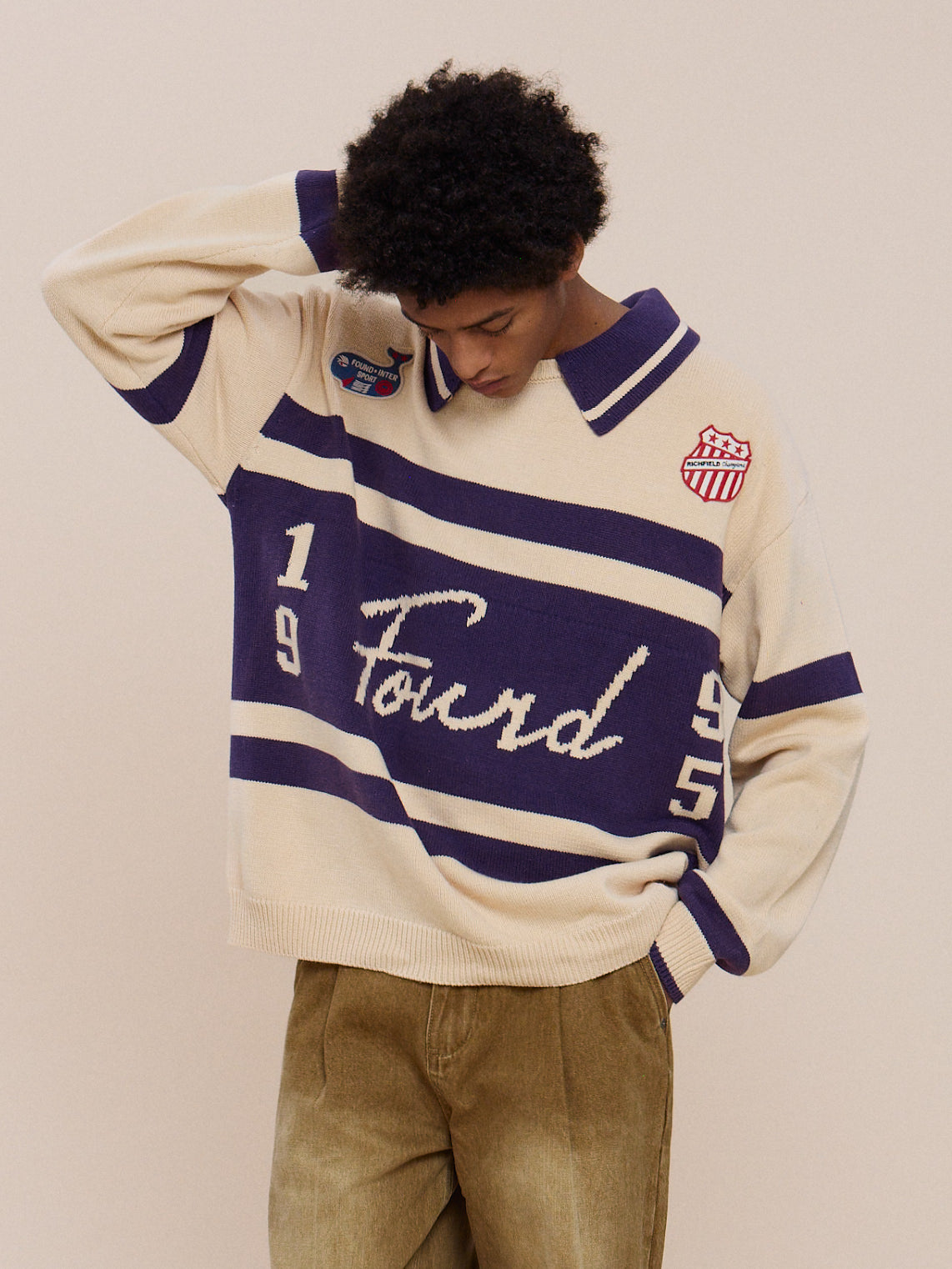 A person in the Knit Jersey by FOUND, featuring cream and navy colors with “Found,” “1955,” and two embroidered patches, stands looking down with one hand behind their head.