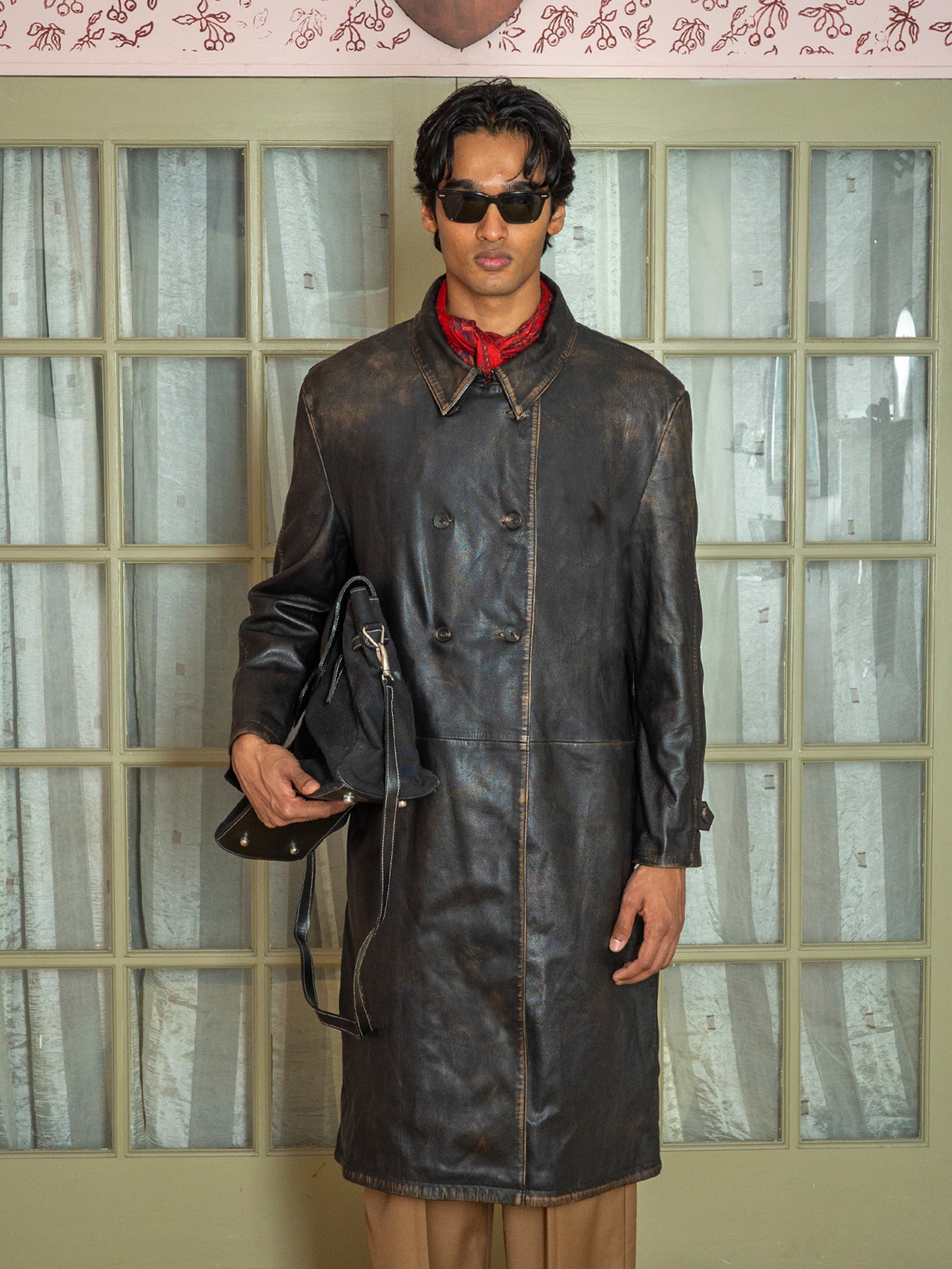 A man in a FOUND Distressed Leather Trench Coat, sunglasses, and a red scarf holds a black bag while standing before glass doors with white curtains.
