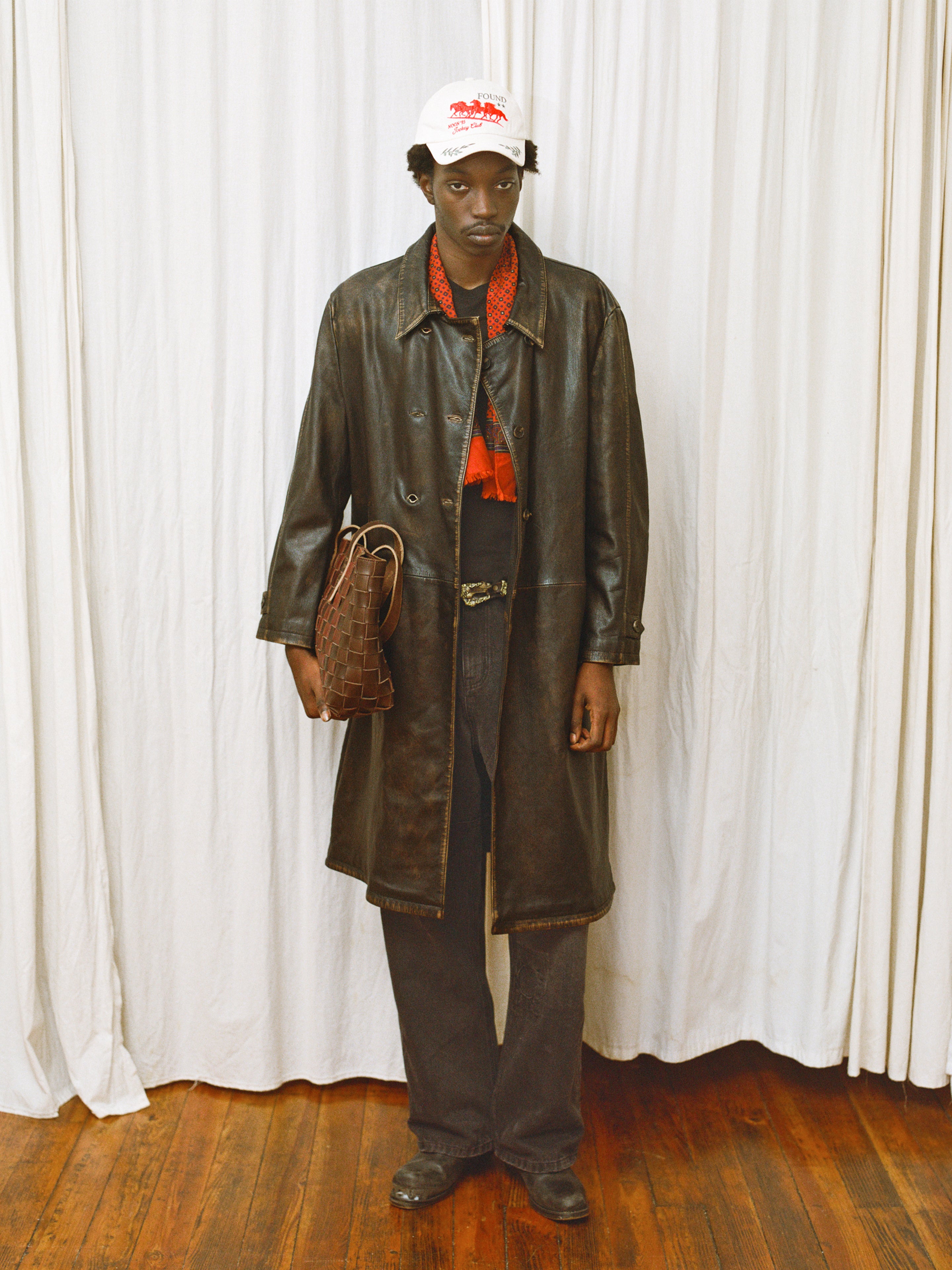 Indoors on a wooden floor, a person wears the FOUND Distressed Leather Trench Coat with black pants, red scarf, white cap, and holds a brown woven bag; a white curtain hangs in the background.