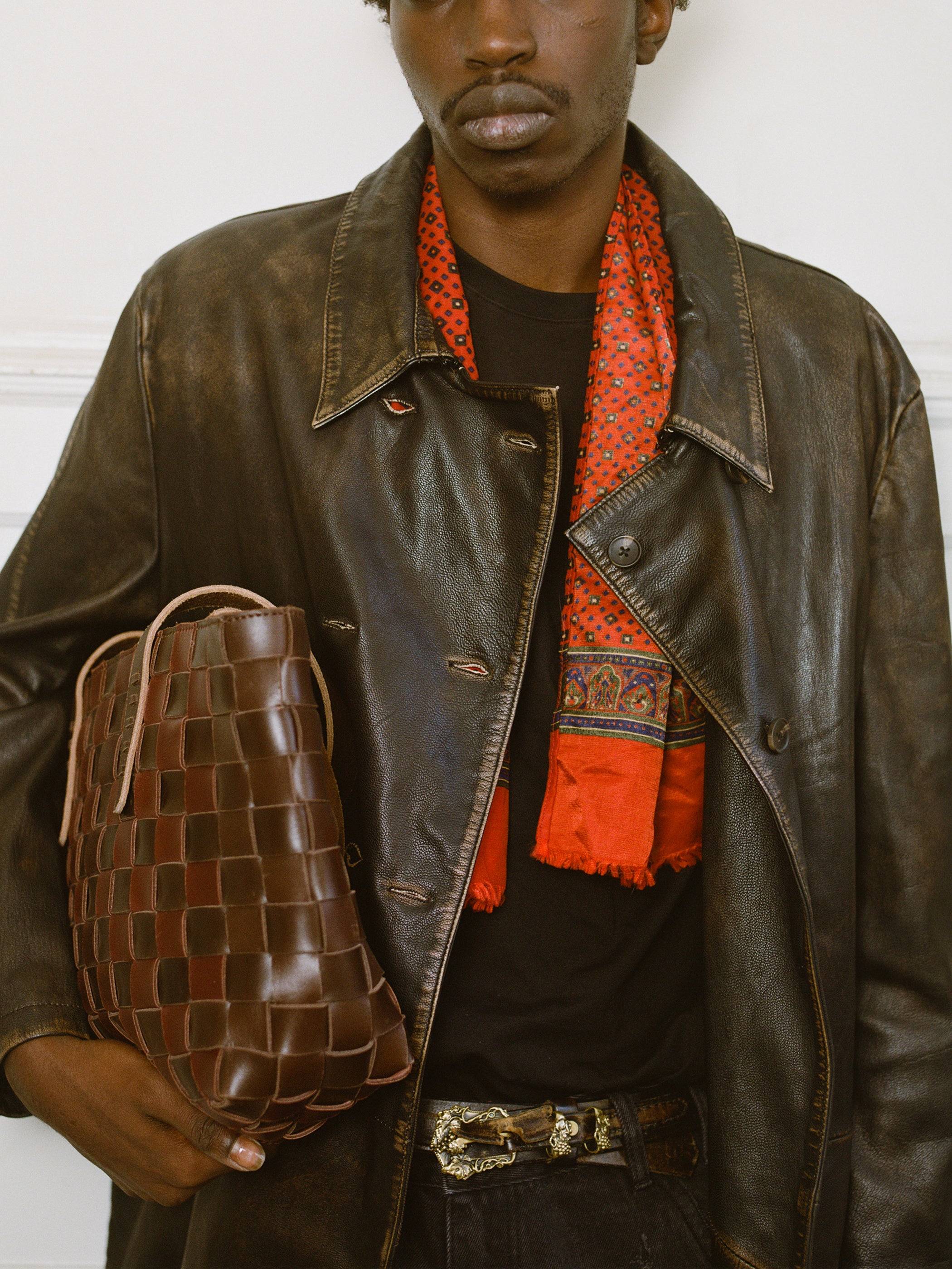 A person wearing the FOUND Distressed Leather Trench Coat, a red patterned scarf, and a black shirt holds a brown woven leather bag in one hand.
