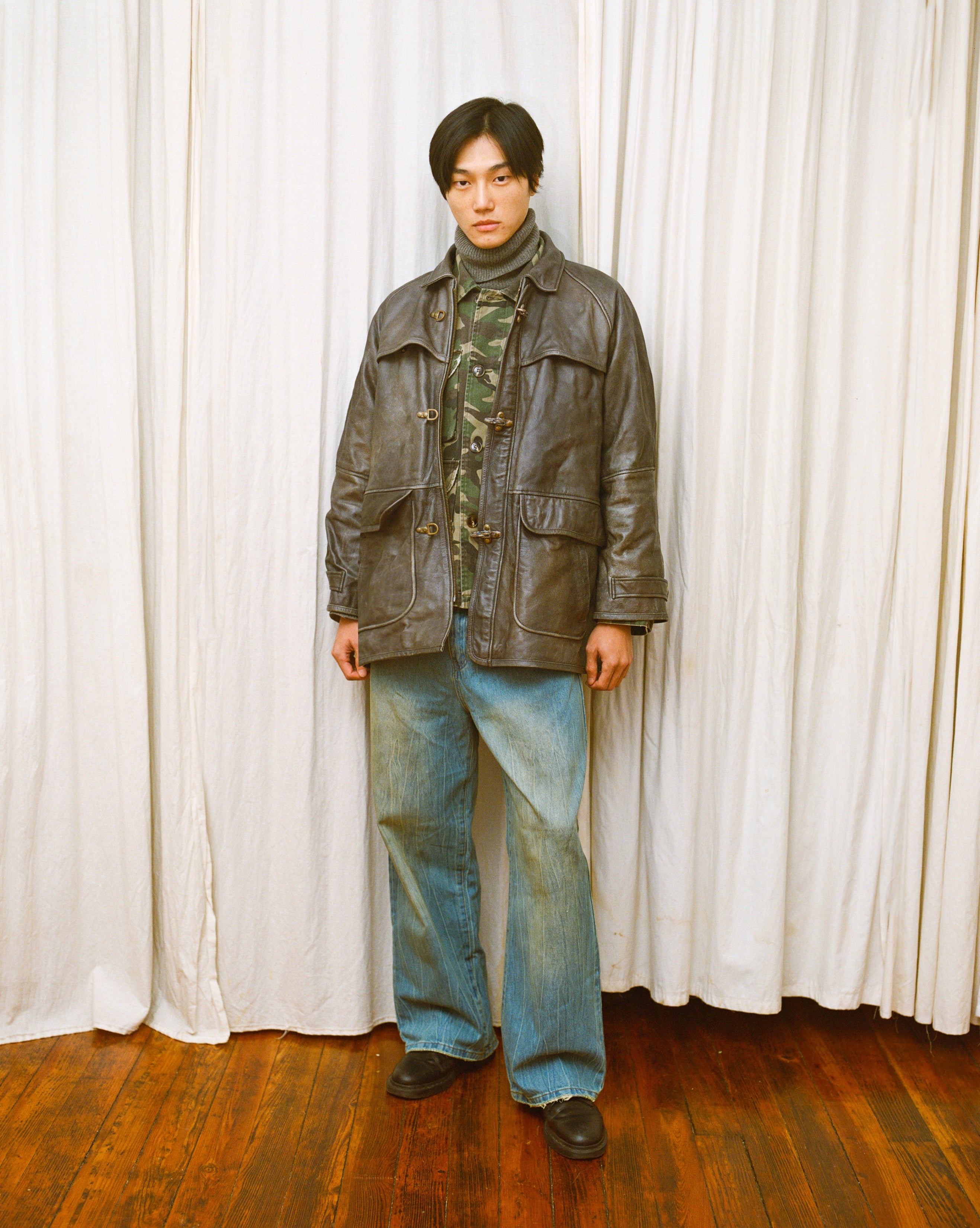 A person stands indoors on wooden flooring, wearing a leather jacket, camouflage shirt, FOUND Lacy Baggy Jeans in dirty blue wash, and dark shoes, in front of white curtains.
