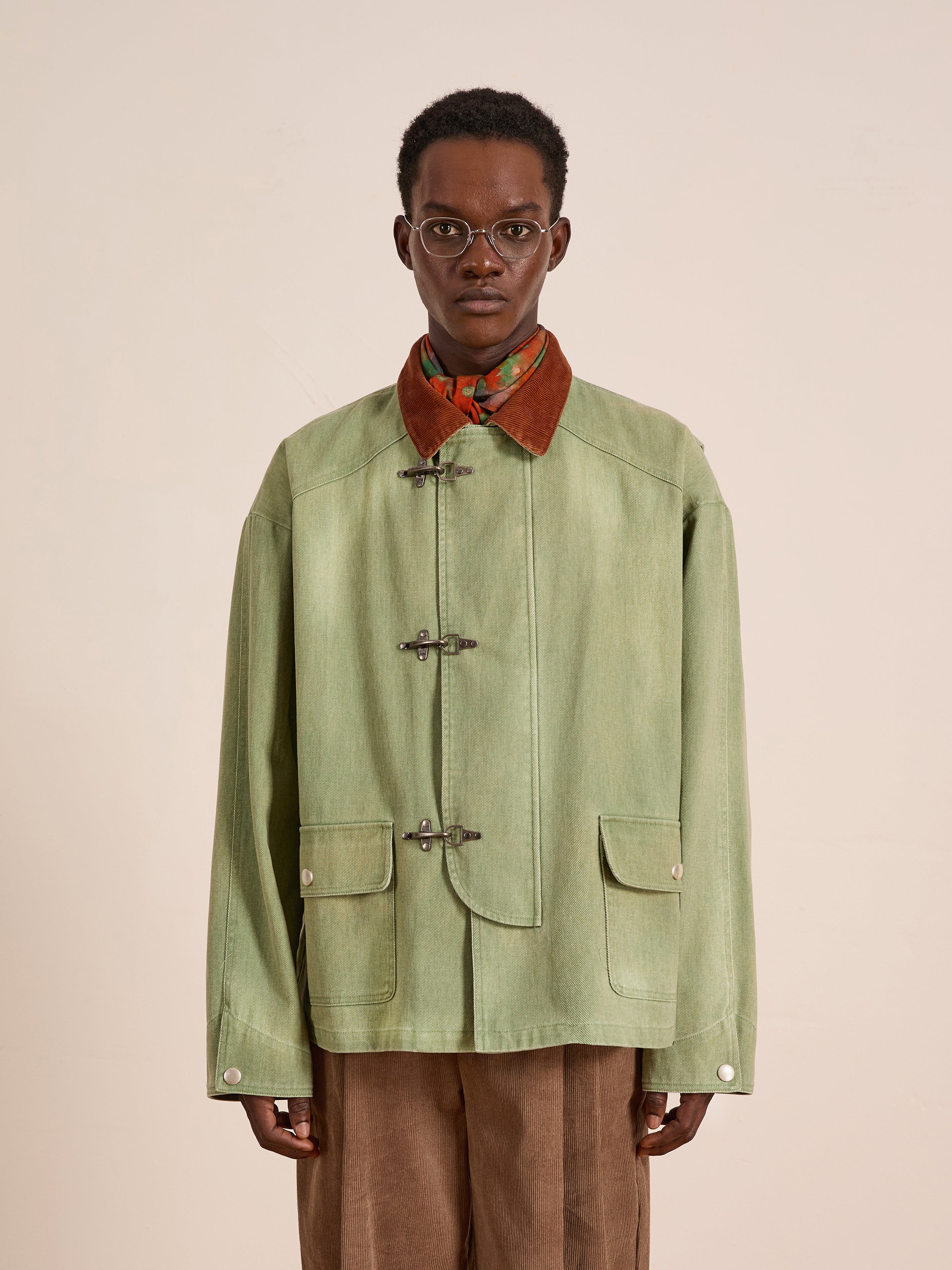 A person stands against a plain background wearing Found’s Washed Fireman Clasp Workshop Jacket, a light green unisex utility piece with toggle closures, two front pockets, and a brown corduroy collar, styled with a scarf and round glasses.