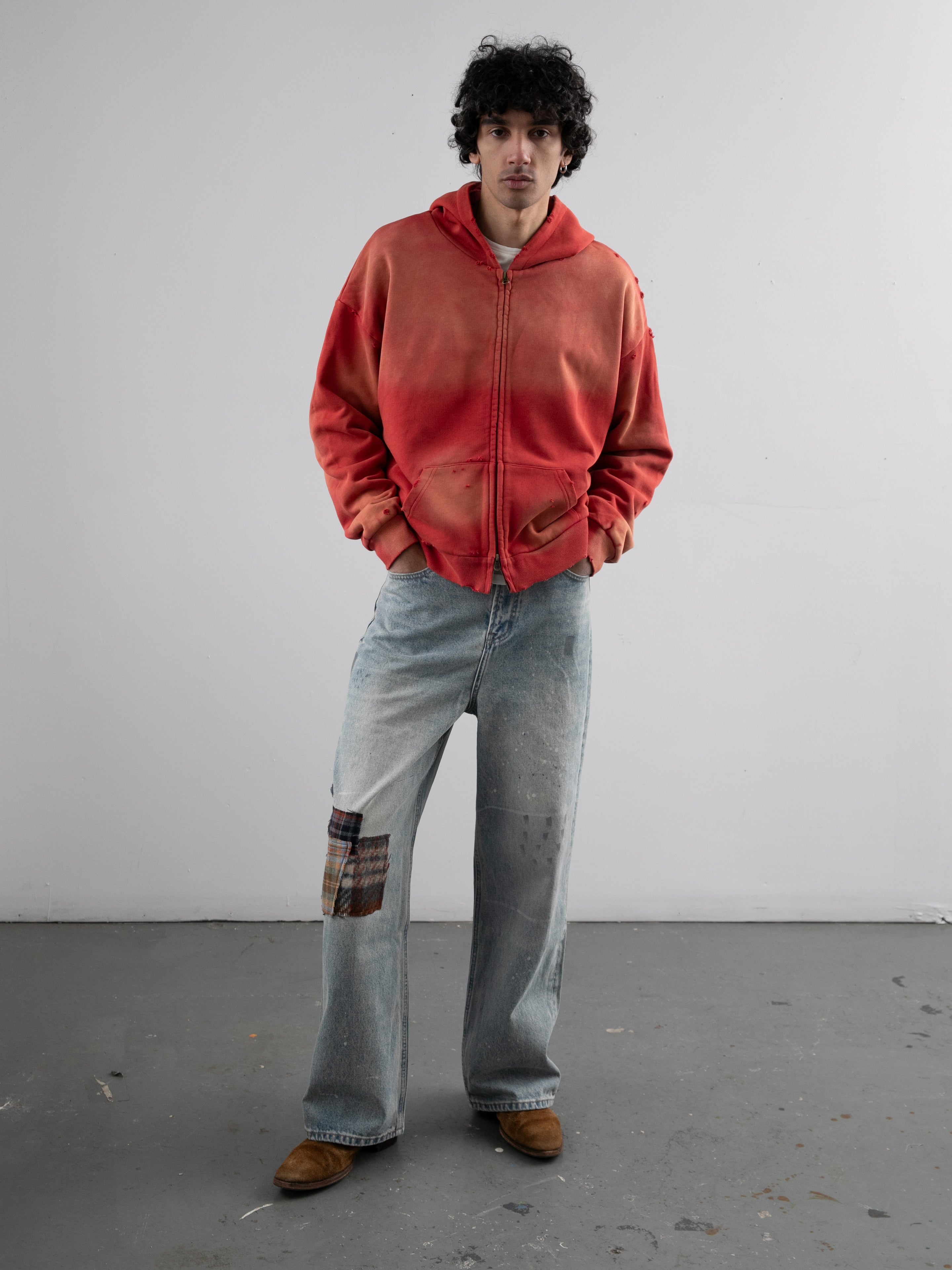 A man stands indoors facing forward with hands in his hoodie pockets, wearing a faded red zip-up hoodie, Profound Mix Patch Jeans, and brown shoes.