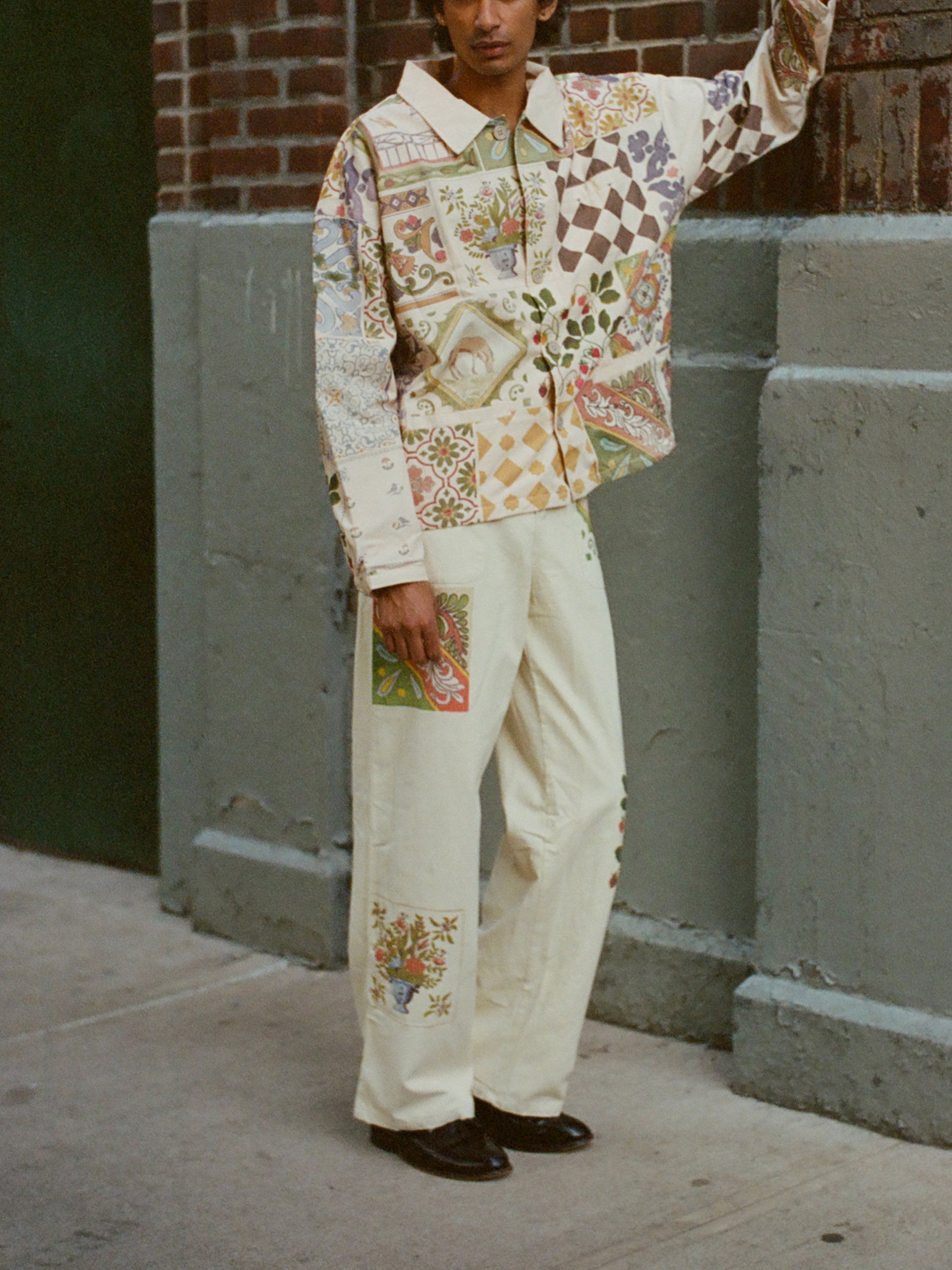Against a brick and concrete wall, someone wears FOUND's Mosaic Pants—a vibrant, unisex ensemble boasting intricate Indo-Aryan patterns in an elaborate patchwork design.