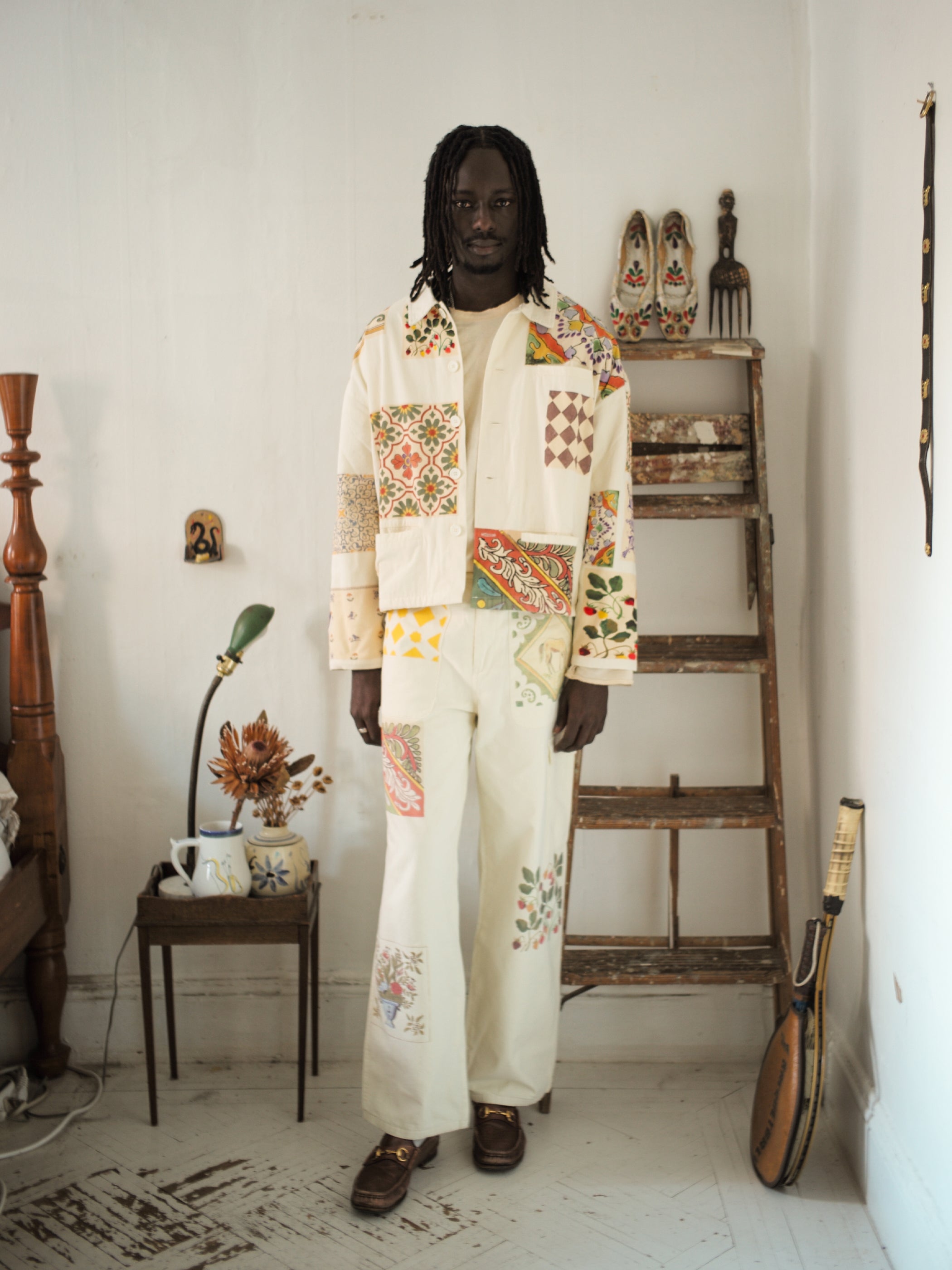 A person stands in a room wearing the cream Mosaic Pants by FOUND, subtly echoing Indo-Aryan patterns. The backdrop features a wooden ladder with decor items and a small table with flowers, complementing the unisex appeal of the outfit.