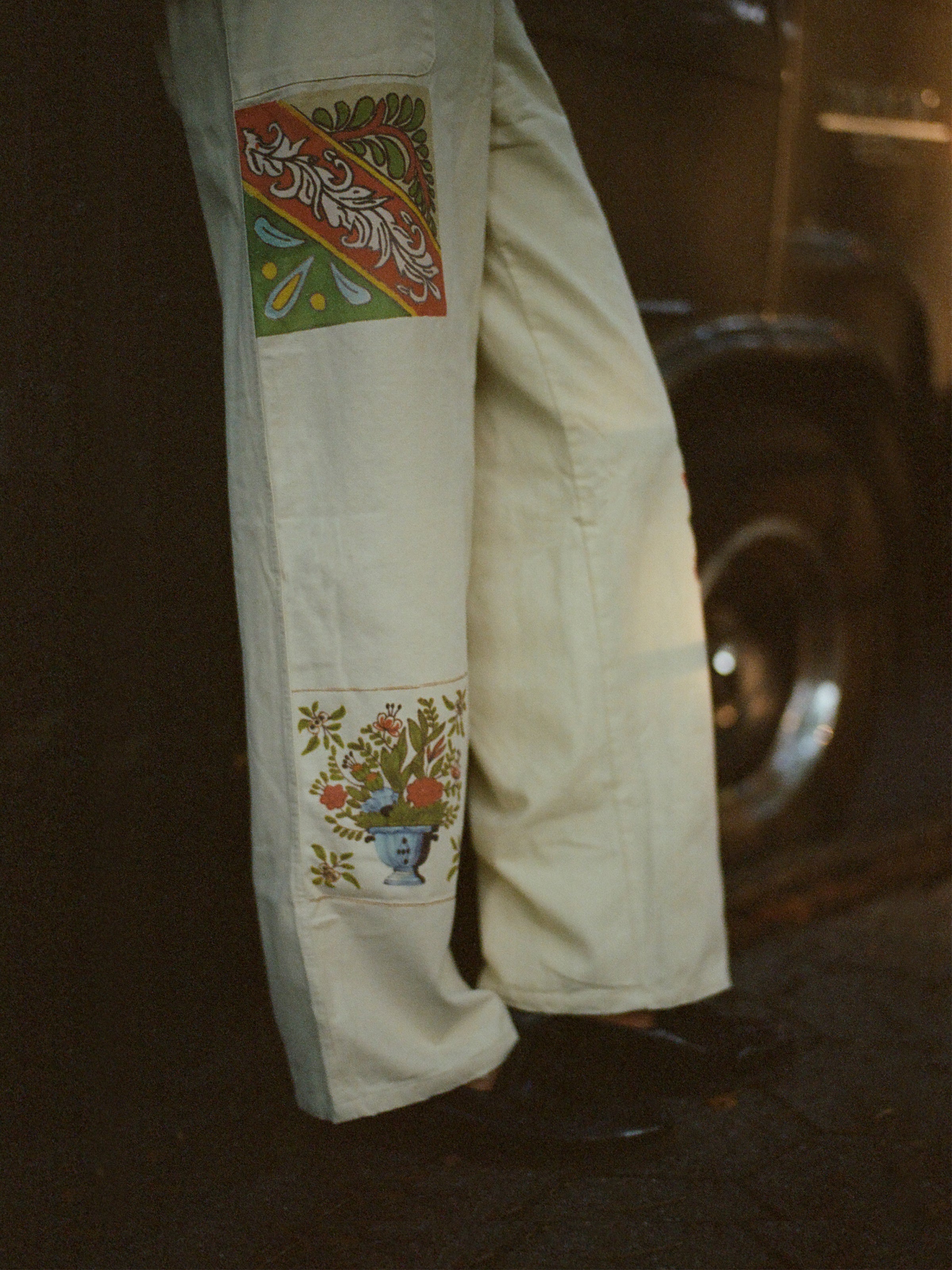 A person stands by a car tire wearing FOUND's Mosaic Pants, featuring cream tones and colorful patches inspired by Indo-Aryan patterns. This unisex design adds versatility to the vibrant look.