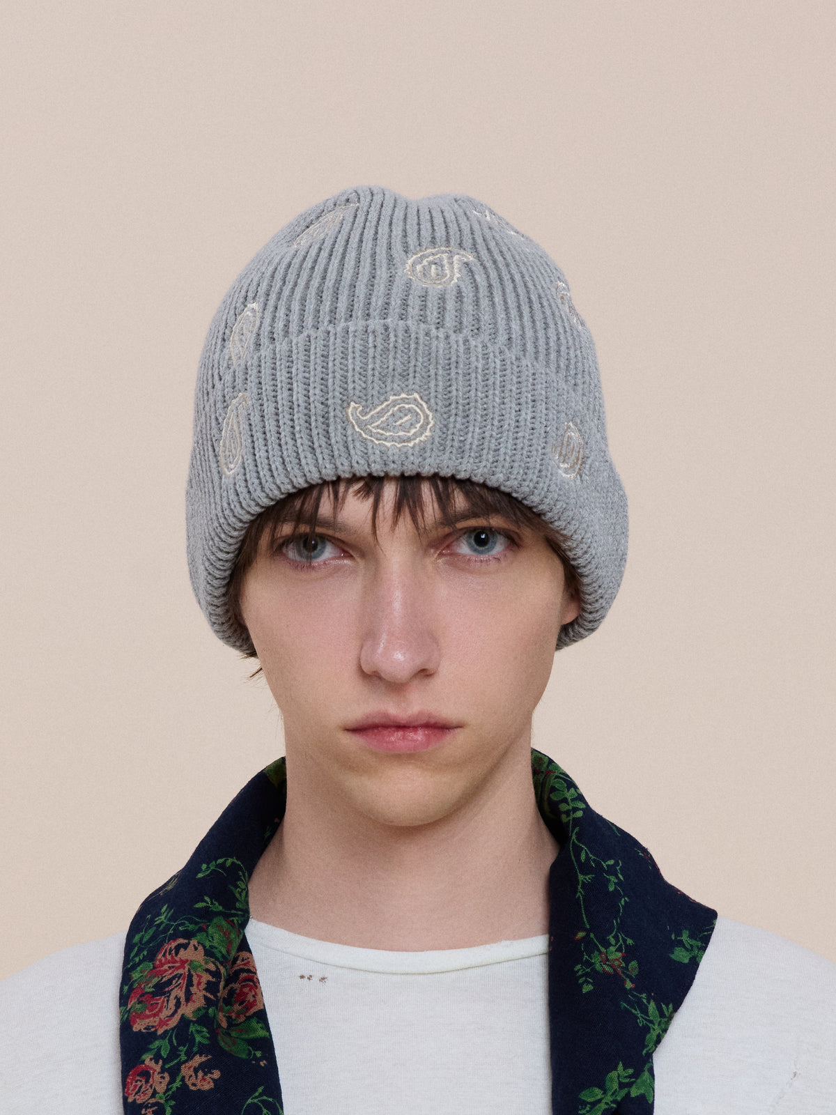 A person with light skin and blue eyes wears a FOUND Paisley Beanie, white shirt, and dark floral-collared jacket, facing forward against a plain background.