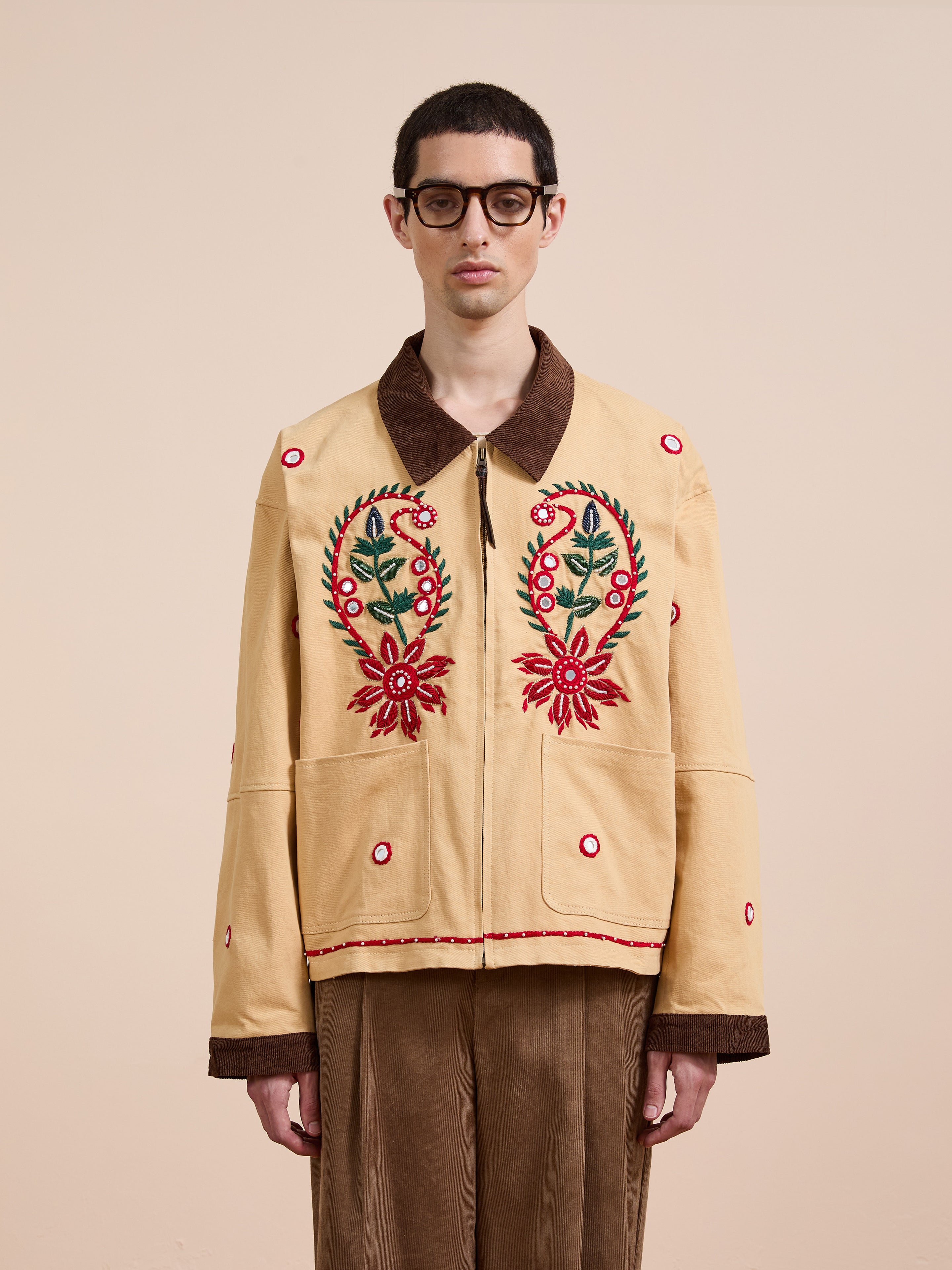 A person wearing glasses and the Found Paisley Motif Hand Embroidered Work Jacket stands against a plain, light background, highlighting its exquisite red floral embroidery and artisan craftsmanship.