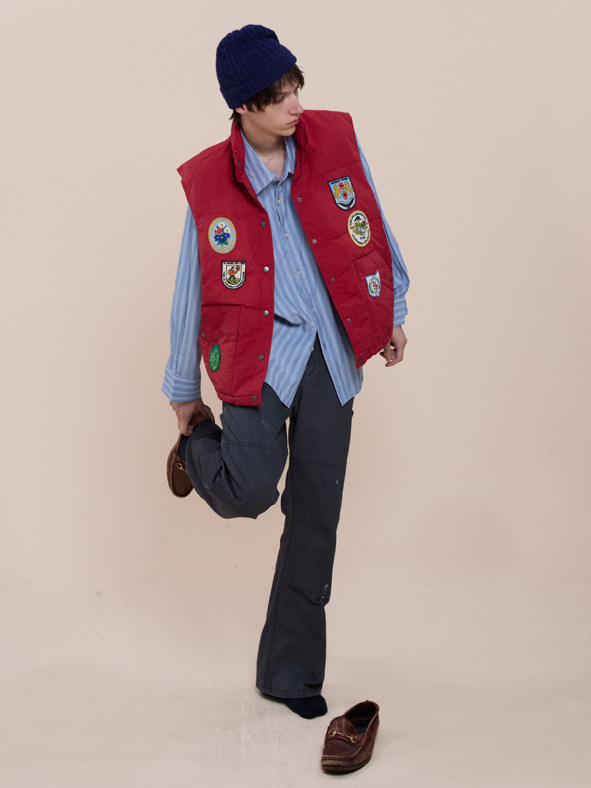 Person in blue beanie, FOUND Patch Down Vest with embroidered patches, blue striped shirt, and dark pants puts on a brown shoe; one shoe lies on the floor. Plain background.