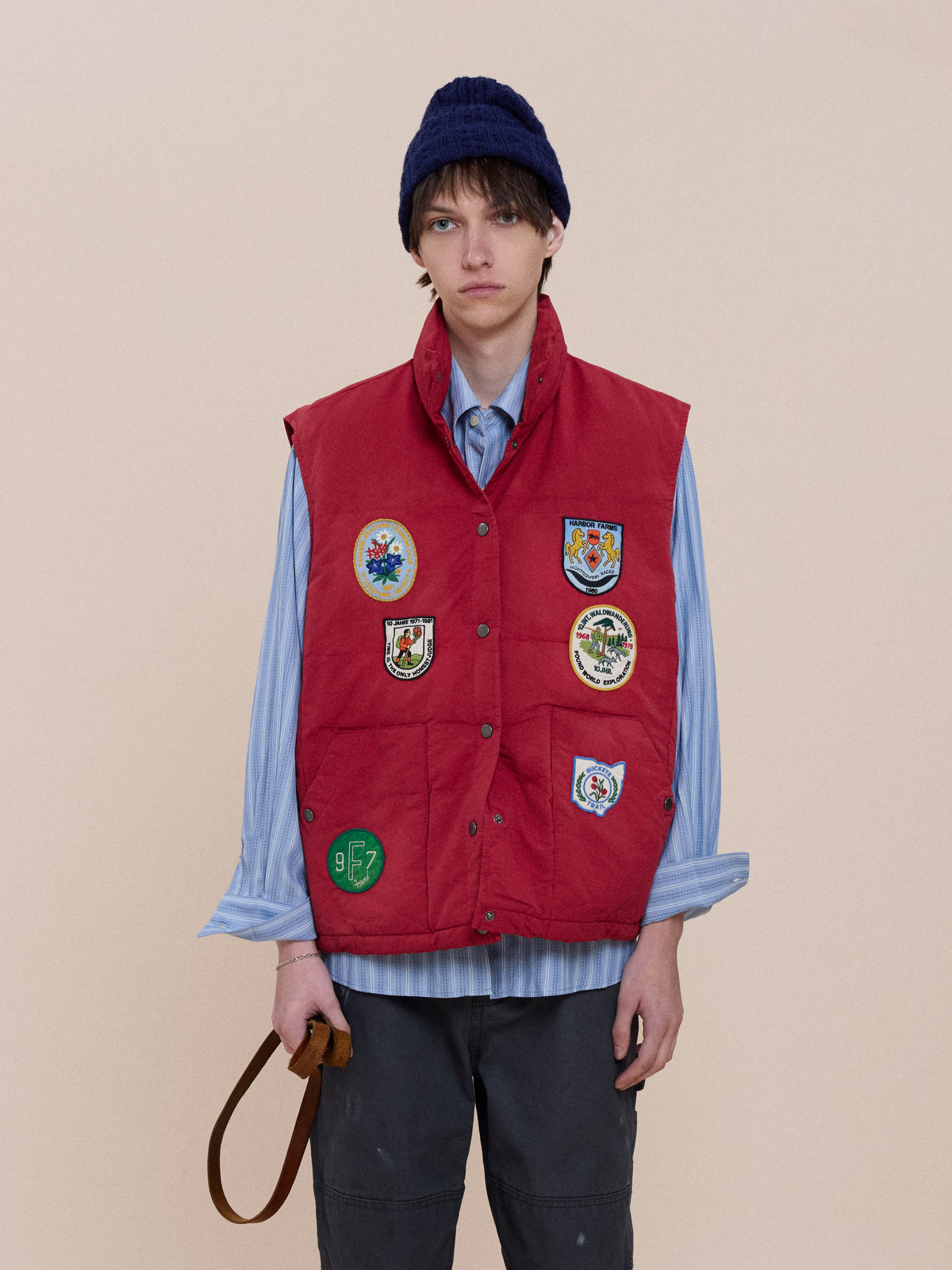 Model wears FOUND Patch Down Vest with embroidered patches, blue knit hat, blue striped shirt, dark pants, and holds a brown strap against a beige background.