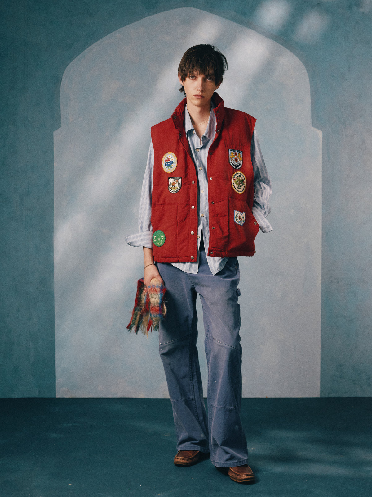 A person models the FOUND Patch Down Vest over a striped shirt and loose blue pants, holding a colorful scarf against an arched blue backdrop.
