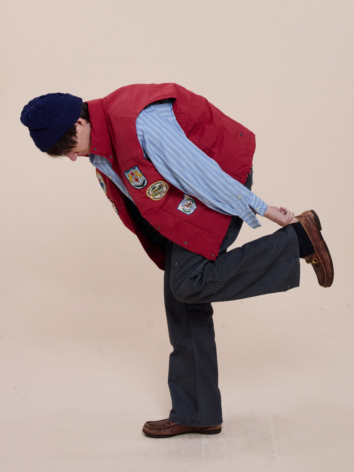 A person wearing the FOUND Patch Down Vest, blue shirt, and dark pants balances on one foot, holding their ankle against a plain background.
