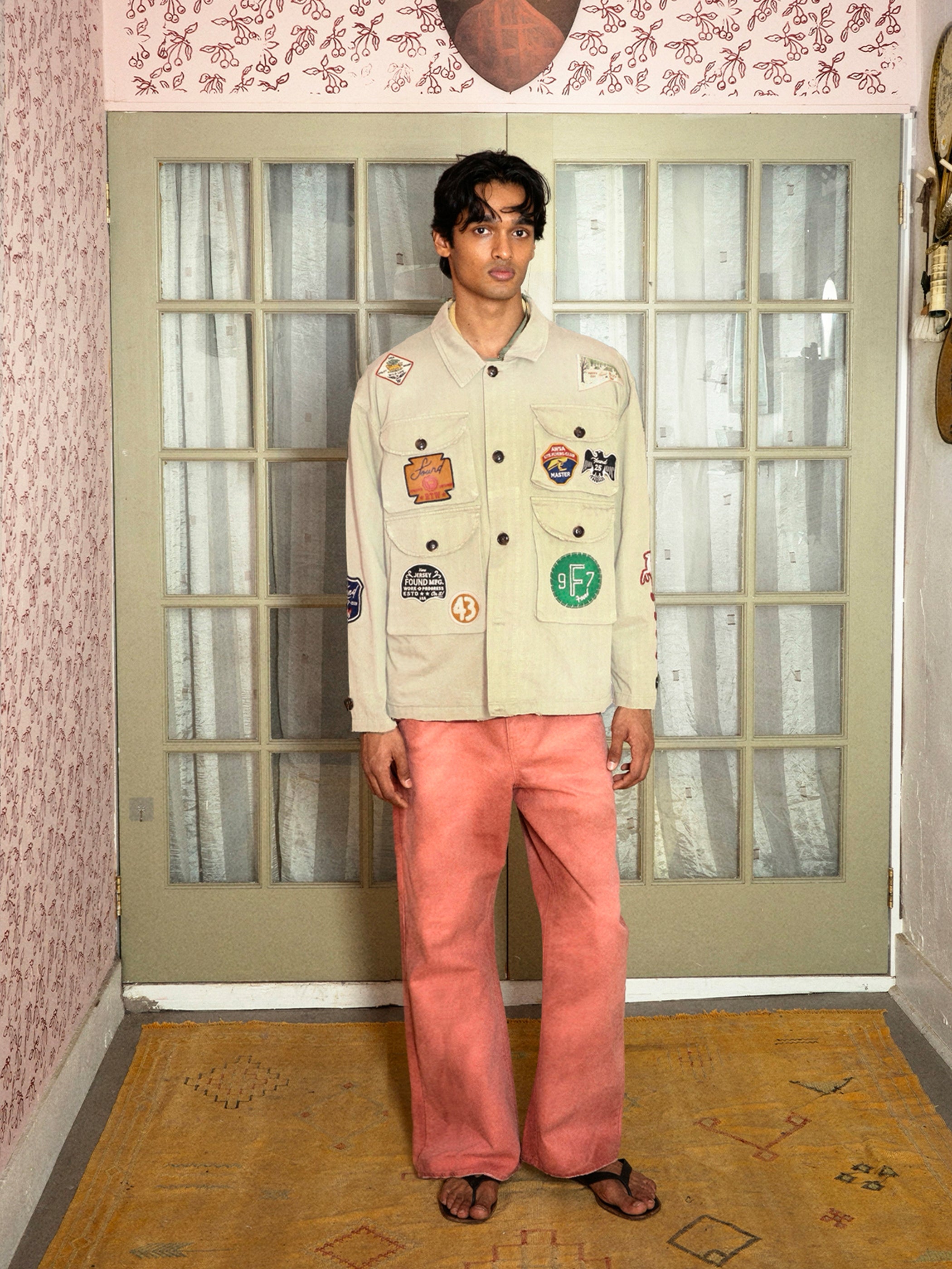 Indoors, a person wears FOUND’s Herringbone Utility Patchwork Jacket with various patches, paired with pink pants and sandals, standing before glass-paned doors draped in sheer white curtains.