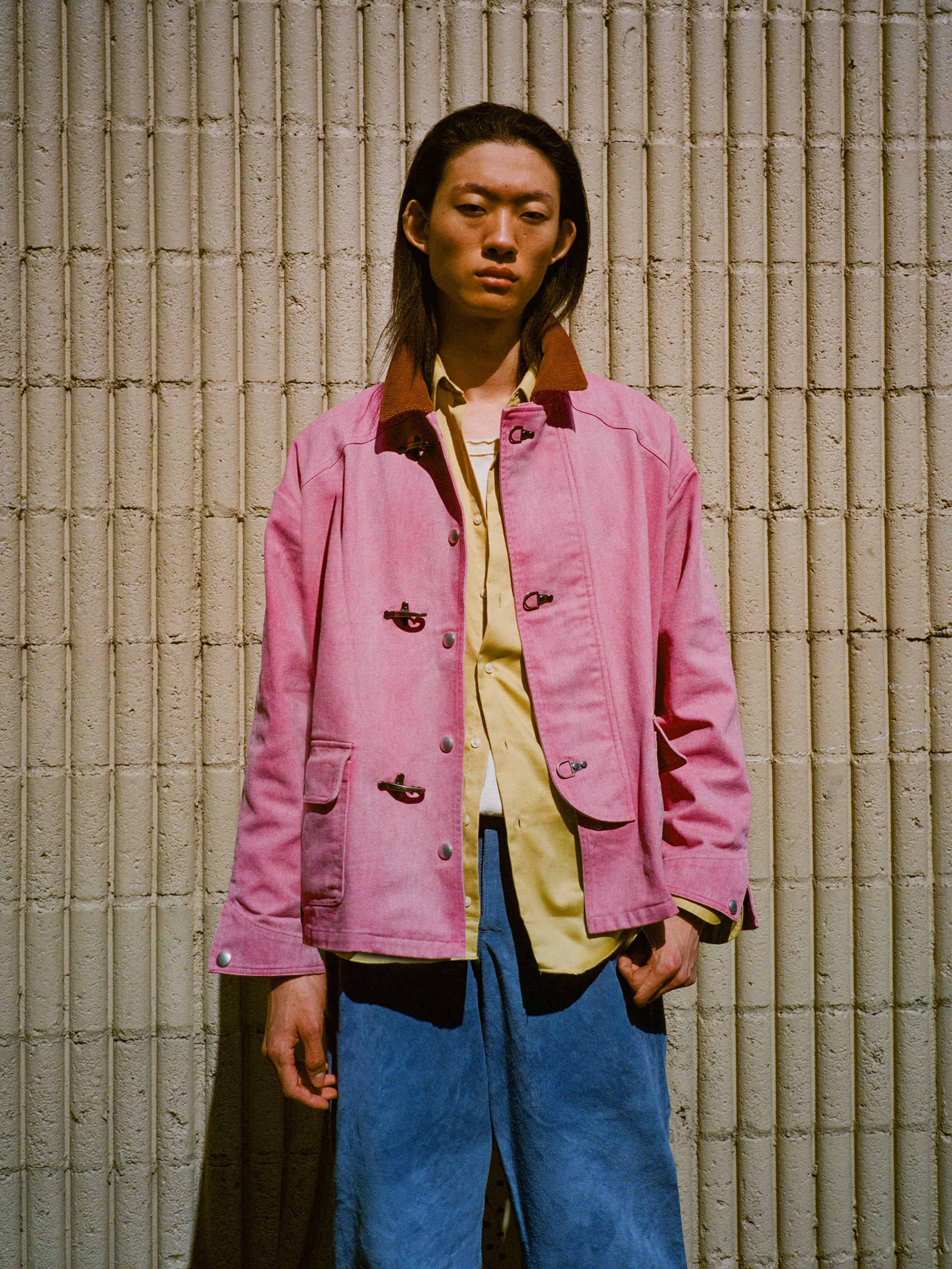 A person stands before a textured beige wall, wearing Found’s Washed Fireman Clasp Workshop Jacket in pink over a light yellow shirt and blue pants.