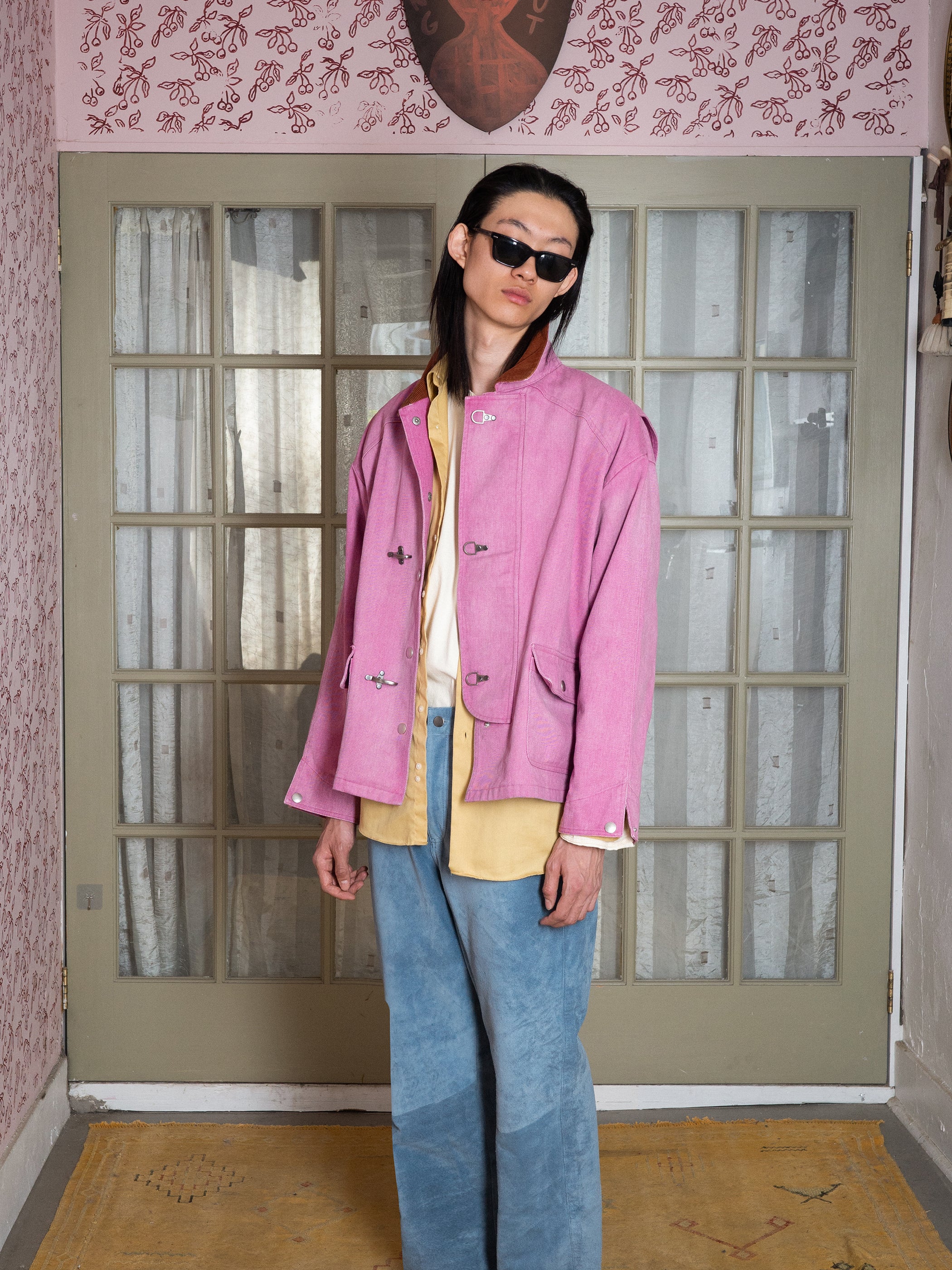 Someone is wearing Found's Washed Fireman Clasp Workshop Jacket over a yellow shirt, with blue pants and sunglasses, standing indoors by glass-paneled doors and patterned pink wallpaper.
