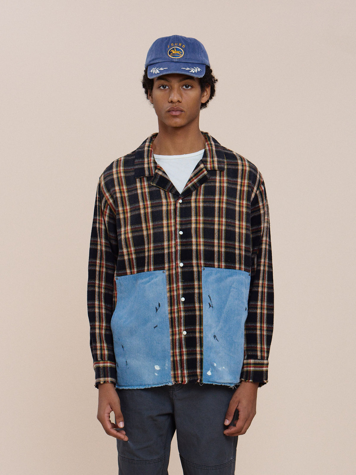 A person in a blue cap, FOUND Plaid Denim Shirt, white undershirt, and dark shorts stands against a plain beige background.