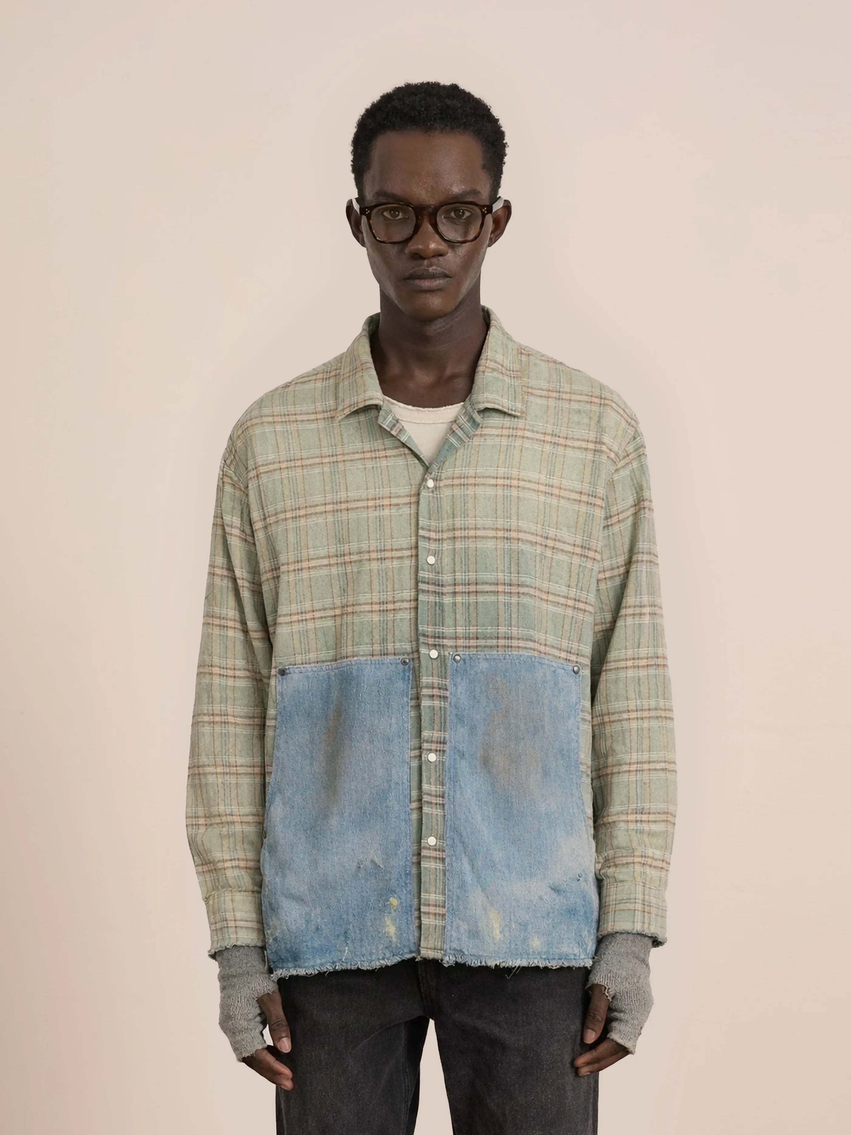A man in glasses stands against a plain background, wearing the FOUND Plaid Denim Shirt—a unisex relaxed fit design with green plaid on top, blue denim fabric on the lower half, and fingerless gloves.