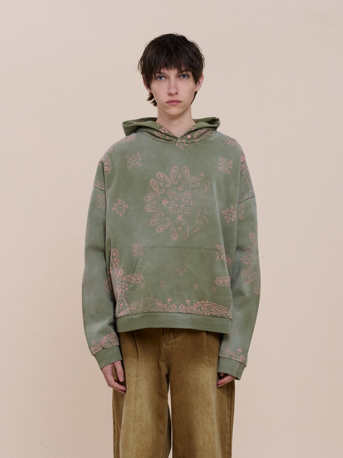 Model wears the FOUND Printed Paisley Hoodie in olive green with tan wide-leg pants, standing against a plain beige background, facing forward.