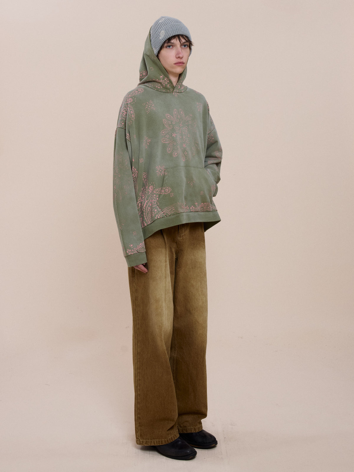 A person wears the FOUND Printed Paisley Hoodie with pink patterns, wide-legged brown pants, black shoes, and a light blue knit beanie in a neutral space.