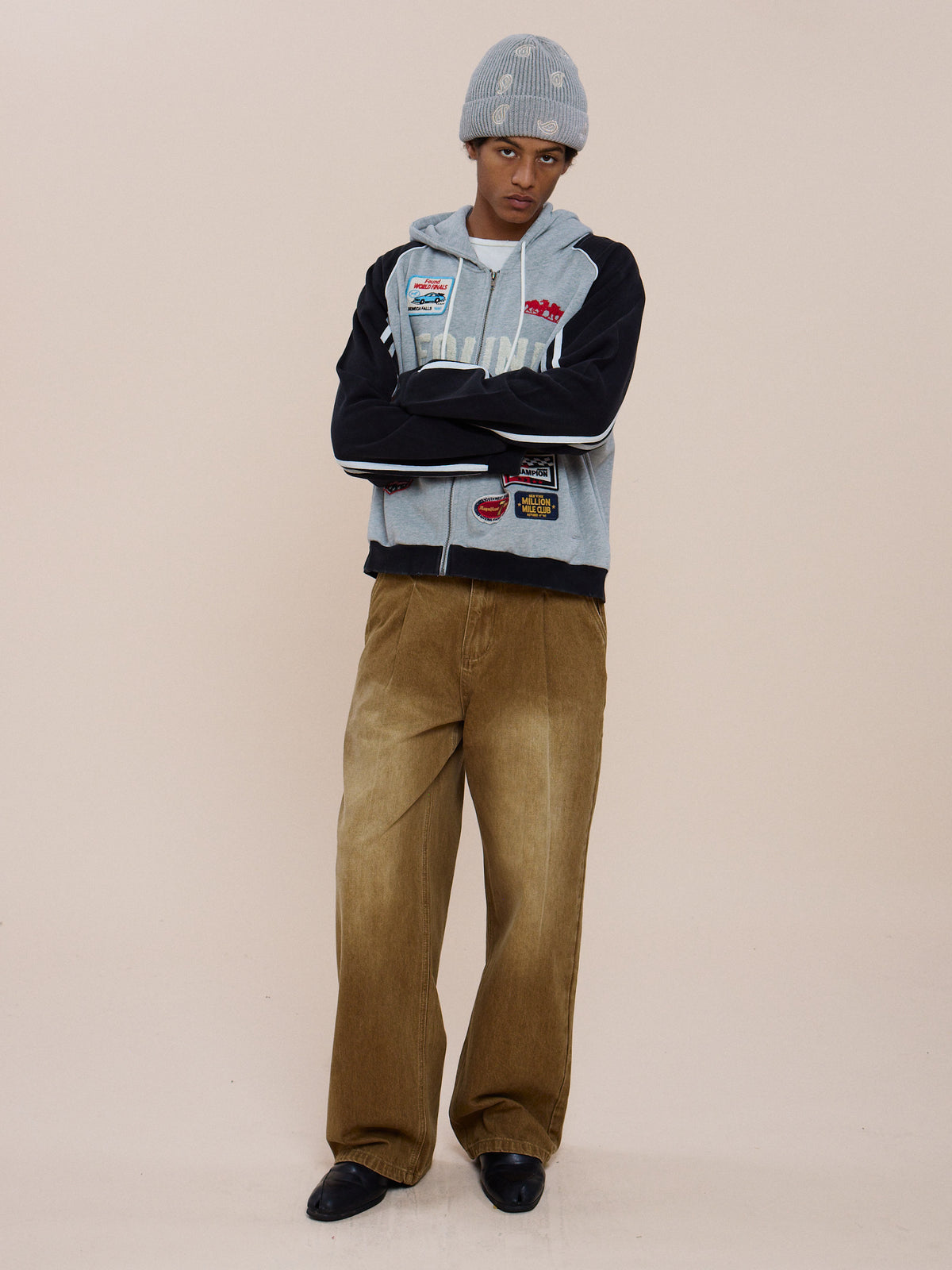 Person standing with arms crossed, wearing a FOUND Racer Hoodie, wide tan corduroy pants, black shoes, and a gray beanie against a plain beige background.