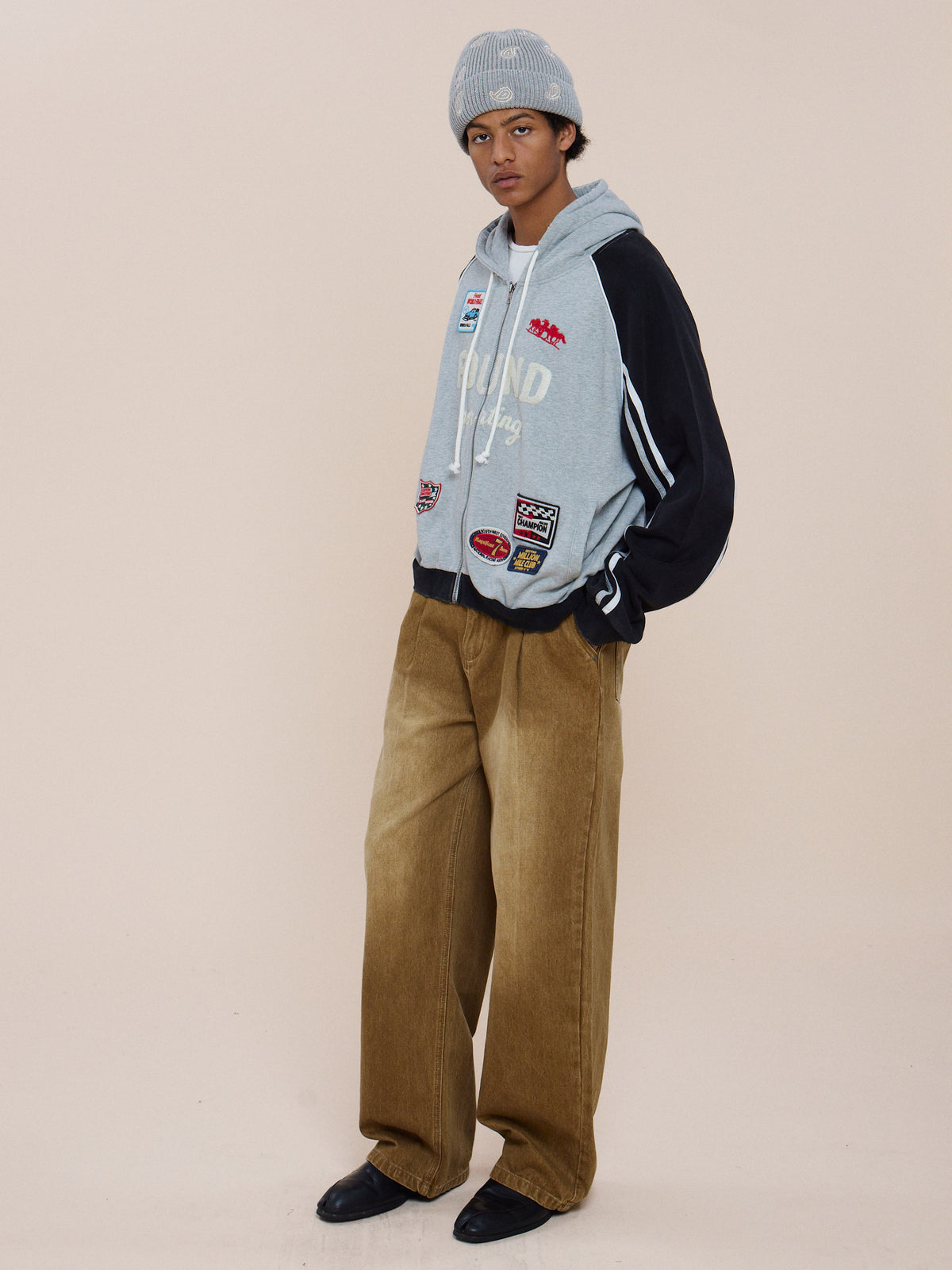A person wears a gray beanie, FOUND Racer Hoodie with patches, wide brown corduroy pants, and black shoes against a plain light-colored background.
