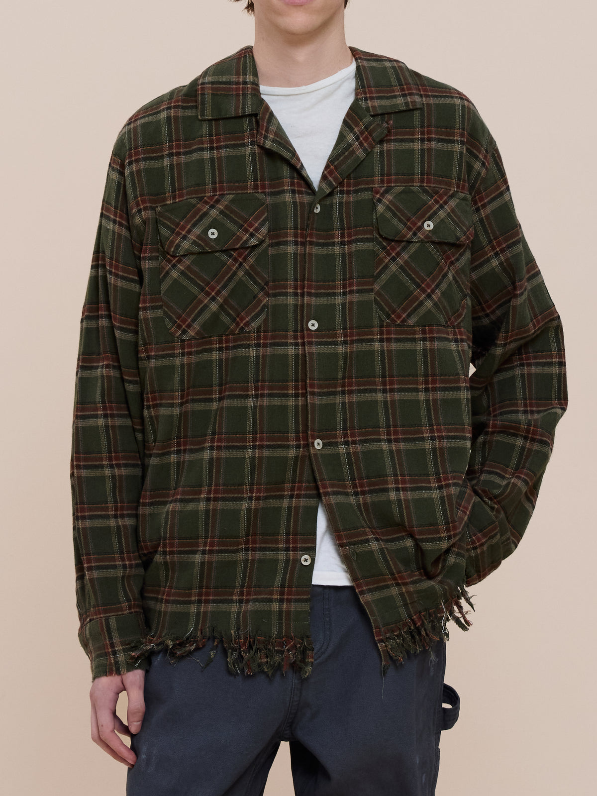 Person wearing the Raw Cut Fringe Plaid Camp Shirt by FOUND over a white tee and dark pants, standing against a plain beige background.