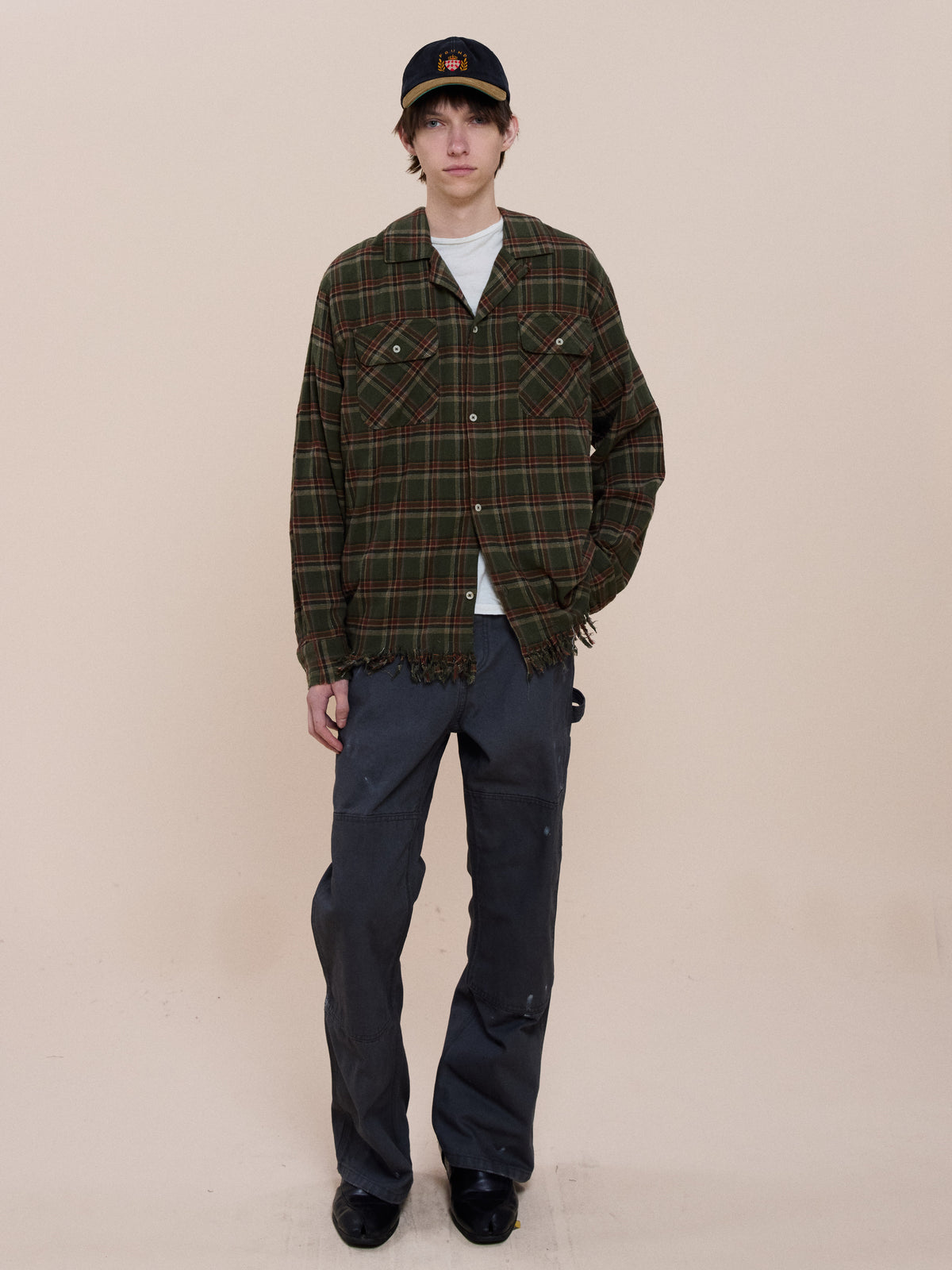 A person in the FOUND Raw Cut Fringe Plaid Camp Shirt over a white tee, dark pants, black shoes, and a black cap stands before a plain beige background.