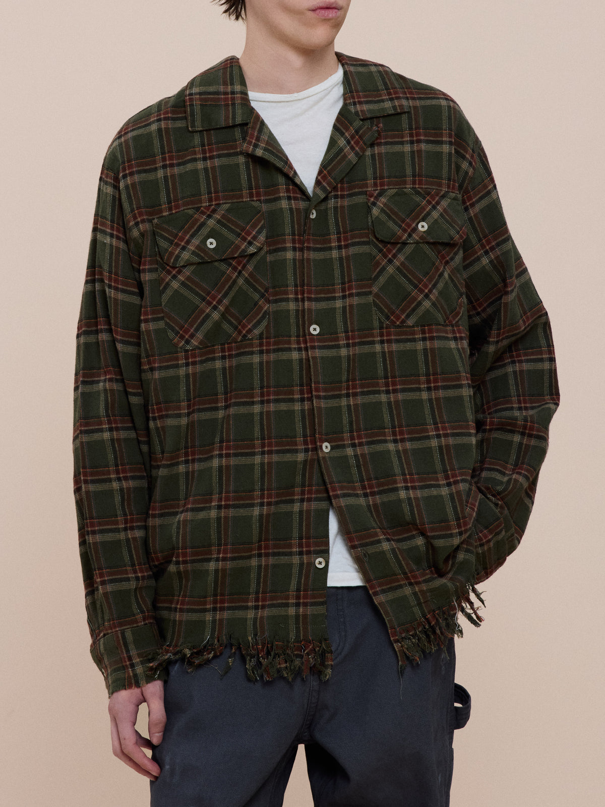 A person wearing the Raw Cut Fringe Plaid Camp Shirt by FOUND over a white t-shirt and dark pants stands against a beige background.