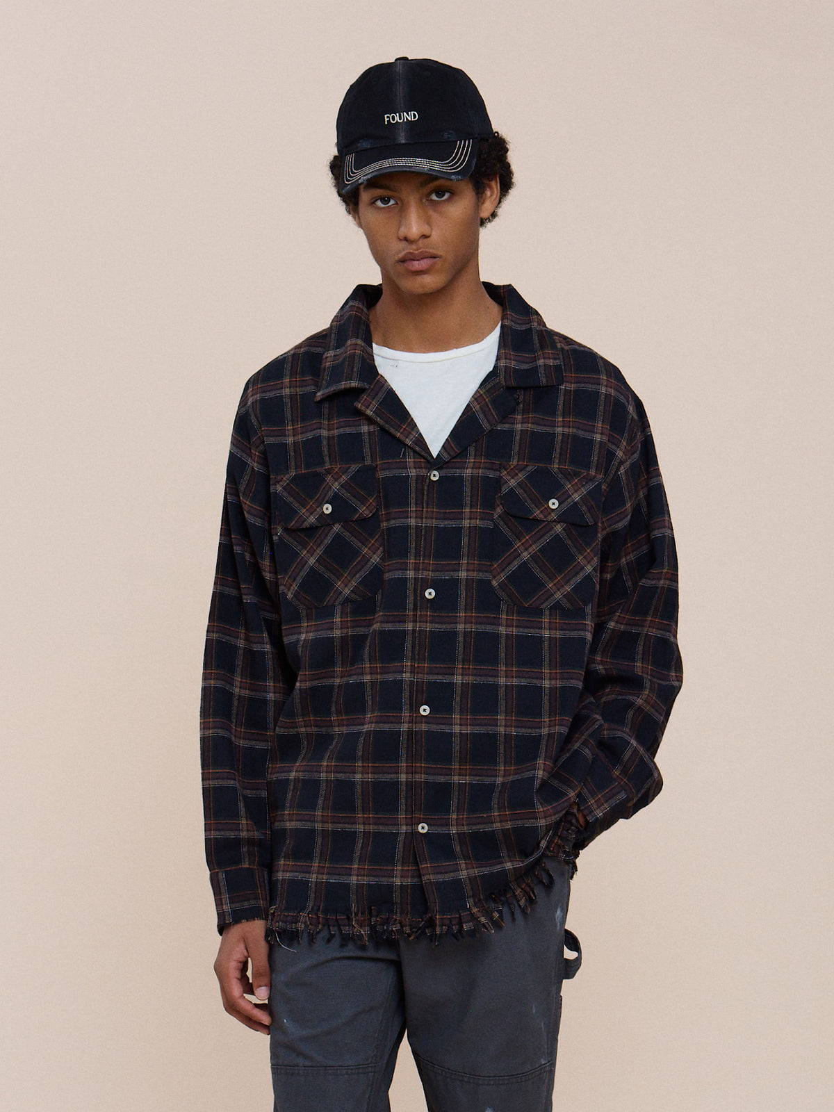 A person wears the Raw Cut Fringe Plaid Camp Shirt, gray pants, and a black FOUND cap, standing against a plain beige background.