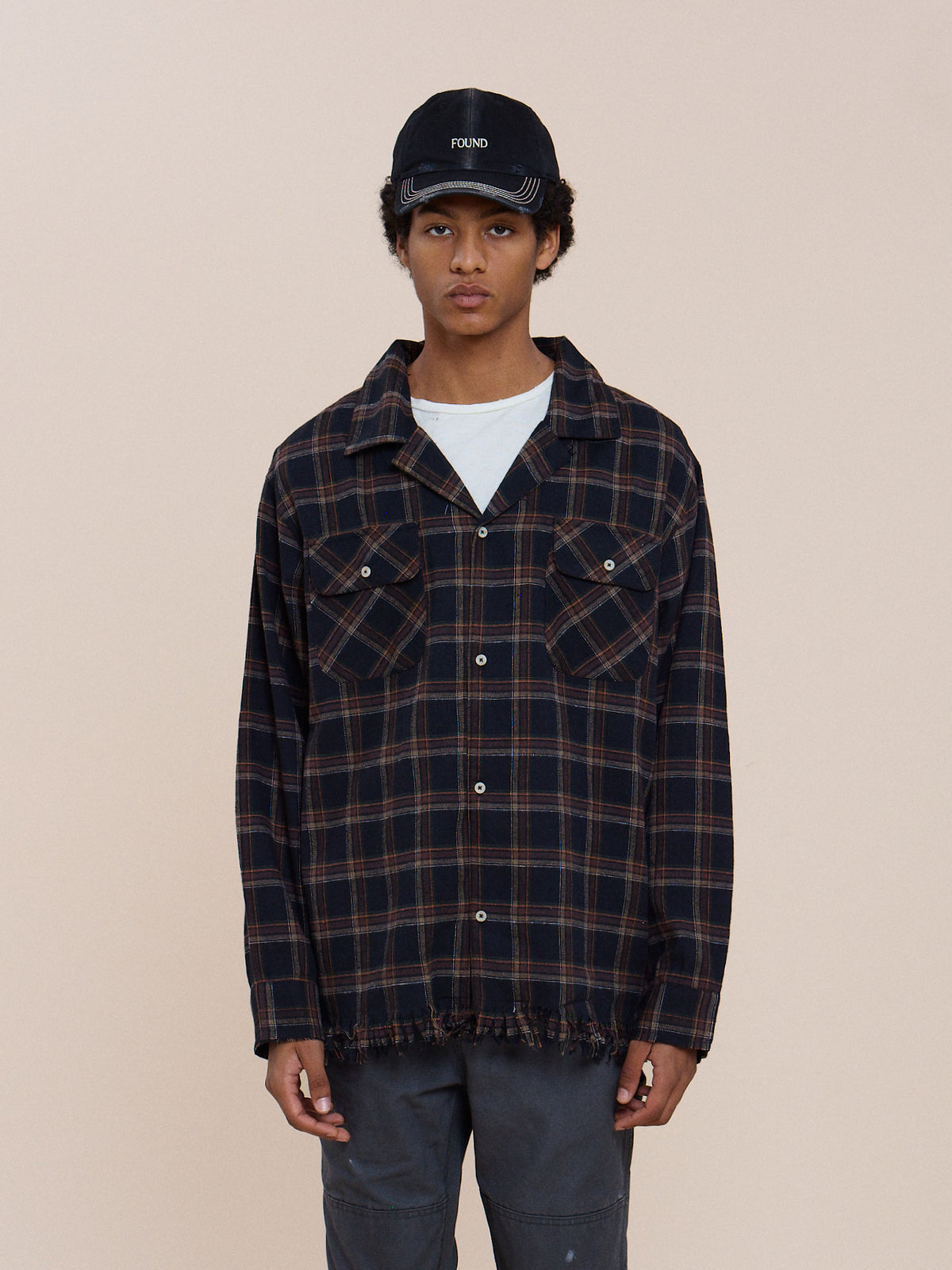 A person stands facing forward in a Raw Cut Fringe Plaid Camp Shirt layered over a white tee, black FOUND cap, and dark pants against a plain beige background.