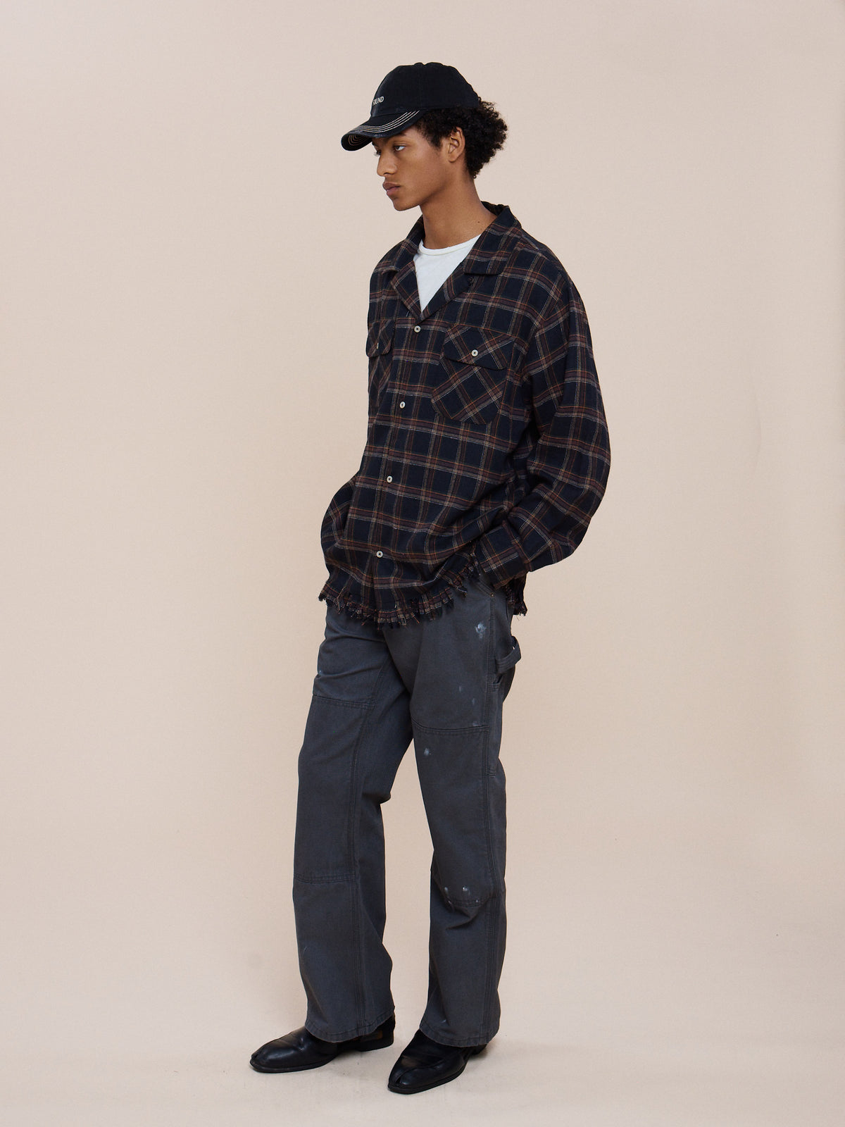 A person in the Raw Cut Fringe Plaid Camp Shirt by FOUND, over a white tee with gray pants, black cap, and black shoes stands hands-in-pockets against a beige background.
