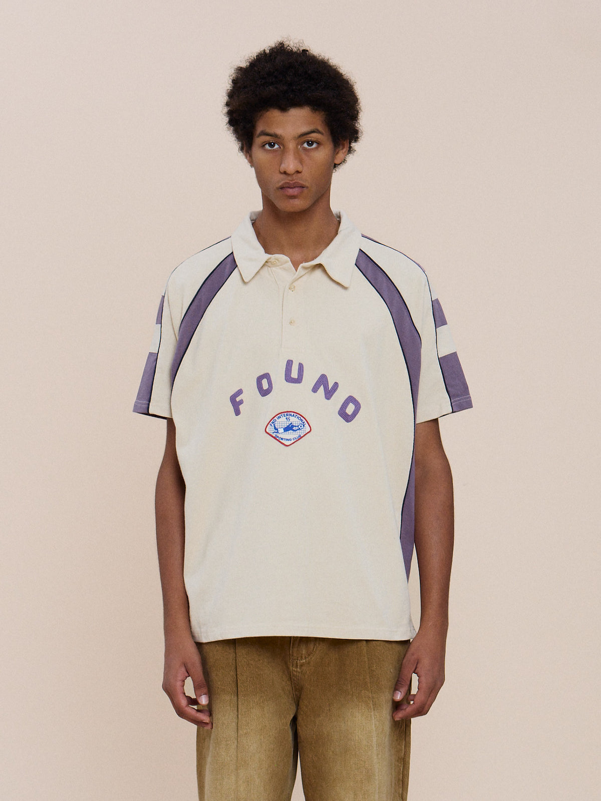 A young person in the Short Sleeve Rugby Polo by FOUND, beige with purple accents and “FOUND” text, relaxed unisex fit, posed against a light background.