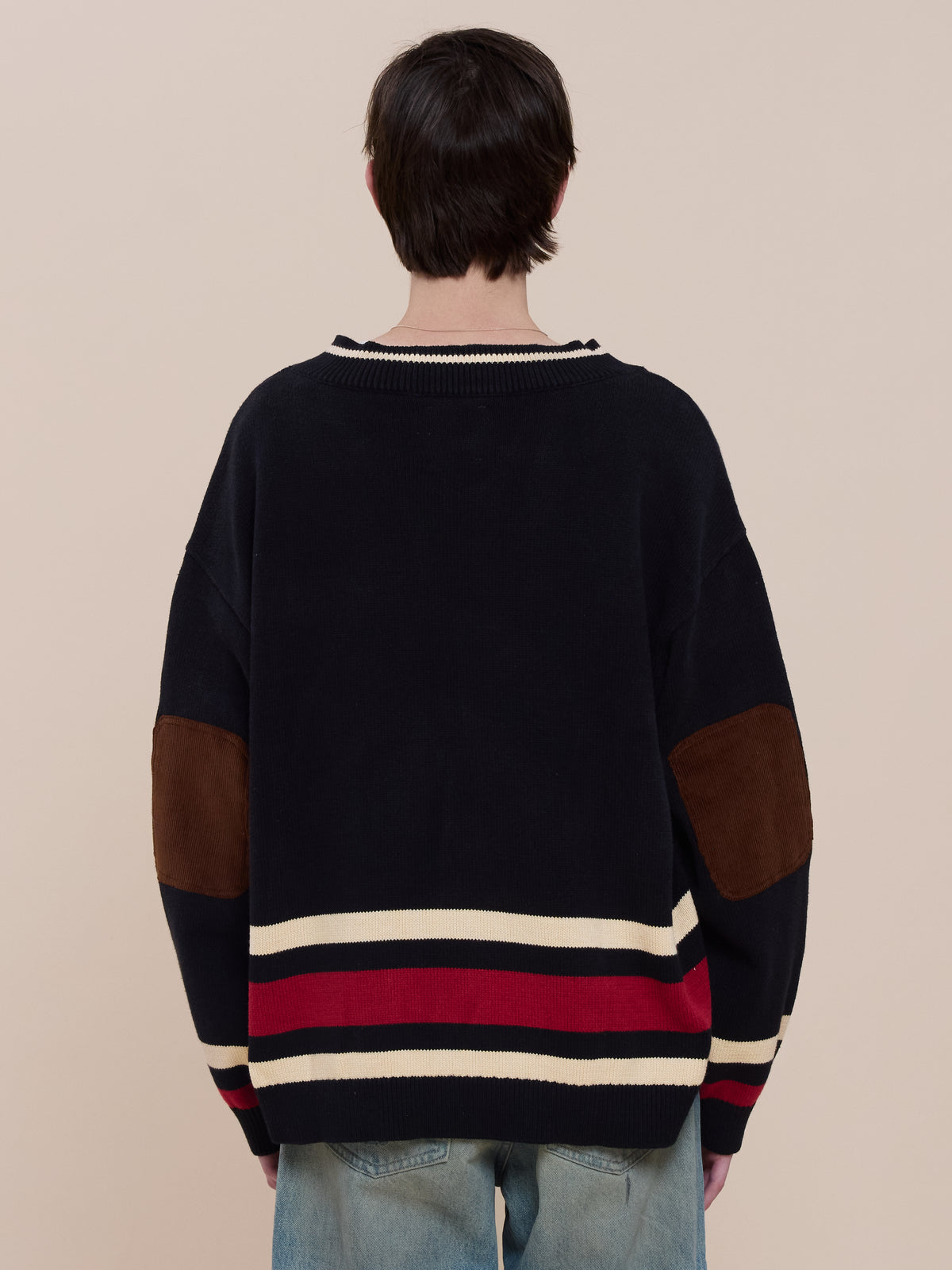 Person standing with their back to the camera, wearing the FOUND Sport Club Varsity Sweater in navy with red and white stripes, brown elbow patches, and light blue jeans.